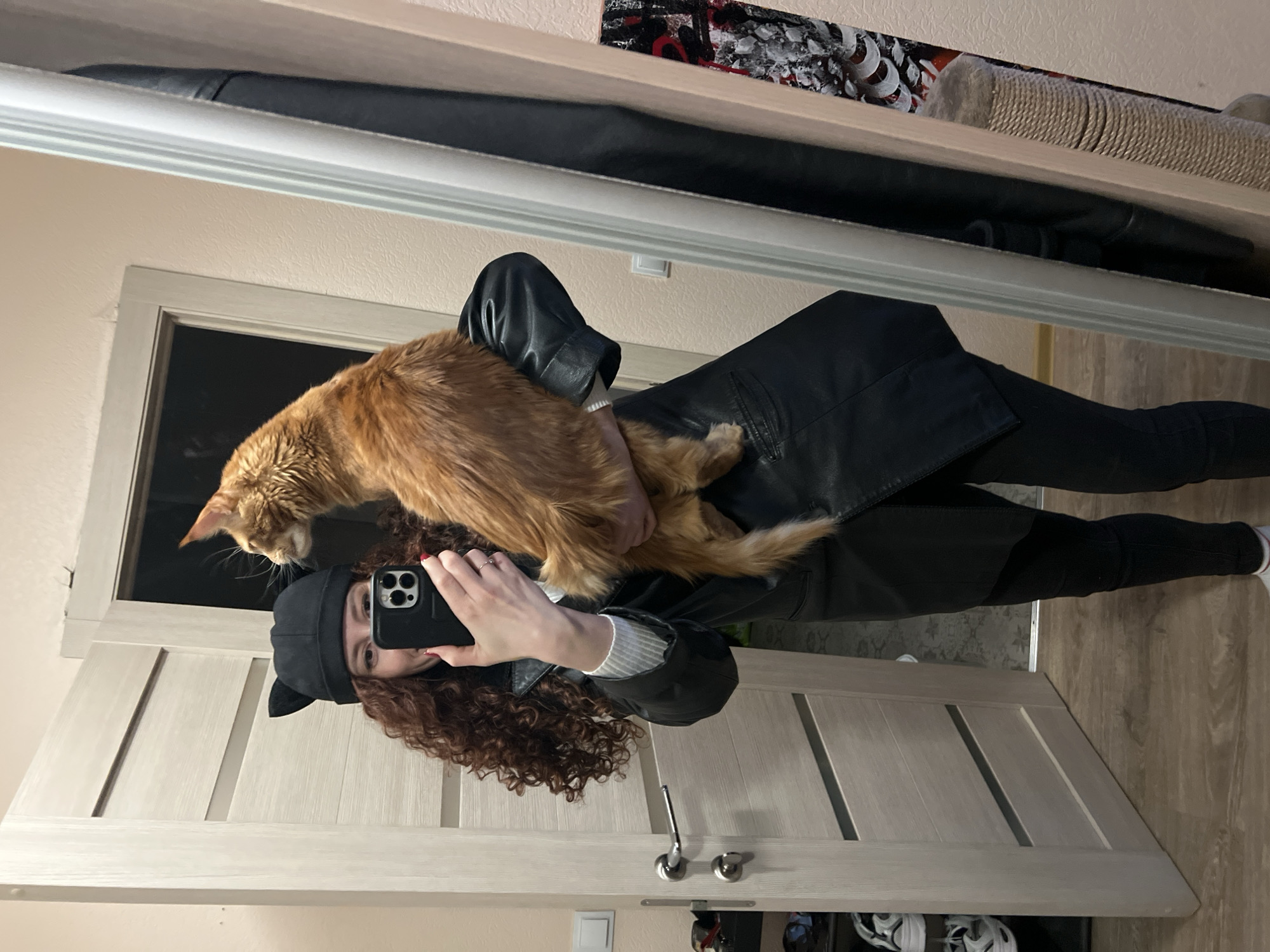 A person is holding an orange cat while taking a selfie in a mirror indoors. The person is wearing a black jacket and black pants, along with a black hat with cat ears. The person is using a black smartphone. A white door and a wall are visible behind the person. Other items are seen on a shelf above the mirror. The location is likely not in the city of Saint Petersburg, Russia.