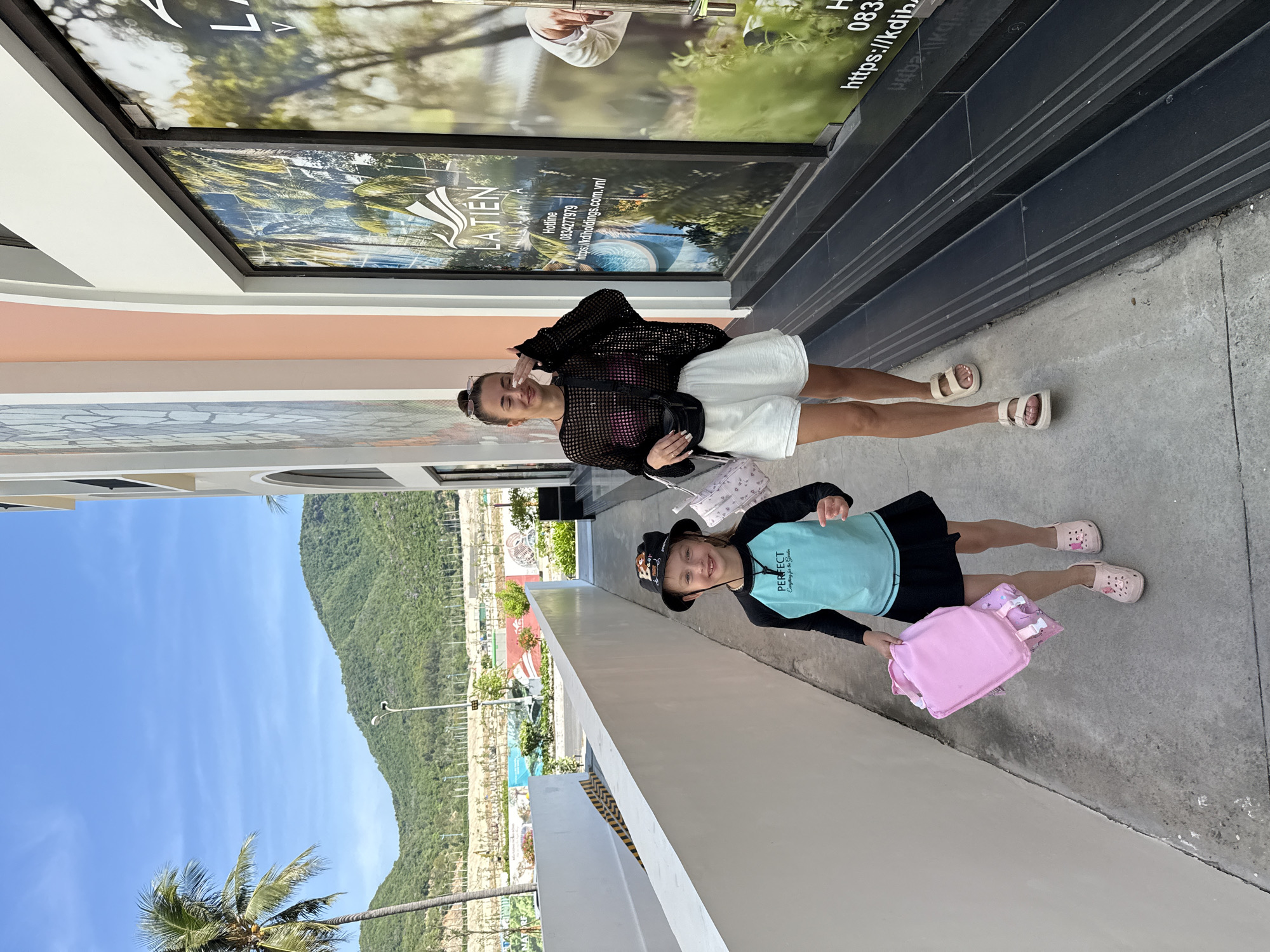 In this eye-level, full-shot photograph taken in Nha Trang, Vietnam, two individuals are posed outside what appears to be a commercial establishment. The scene is illuminated by bright daylight, suggesting it is daytime.
The person in the foreground, a young girl, is wearing a black hat with cartoon characters, a teal long-sleeved shirt with the text "PERFECT" and "Anything for the Garden" printed on it, and a black skirt. She is also wearing pink crocs and carrying a pink bag. Her expression is cheerful and she is looking towards the camera.
Behind her, an adult woman is smiling and making a peace sign. She is dressed in a black mesh long-sleeved top over a pink bikini, white shorts, and light-colored sandals. She has a black fanny pack around her waist and sunglasses on her head.
The background features a large storefront with a banner displaying tropical imagery and text, including "LA TIÊN" and a phone number. Beyond the storefront, a lush green mountainside and a clear blue sky are visible, hinting at a scenic coastal location. The ground is a concrete pathway. The overall atmosphere is casual and summery.
