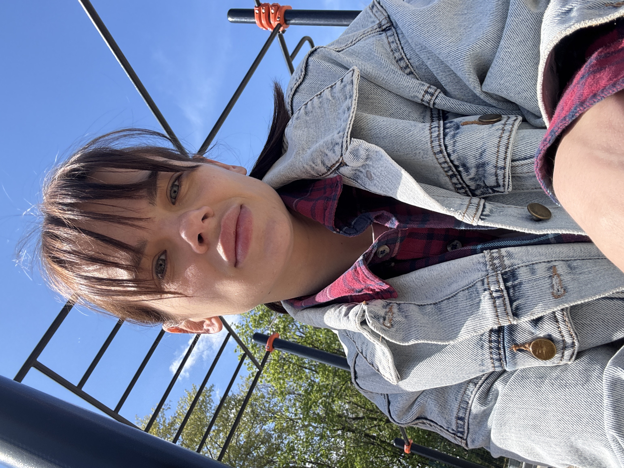 A woman is visible in a low-angle selfie. She has brown hair with bangs, fair skin, and is wearing a denim jacket over a red and black plaid shirt. The background shows a clear blue sky with a metal structure, possibly part of a playground or outdoor gym equipment, with some green trees visible. The specific location in Rostov-on-Don, Russia cannot be determined from the provided image.