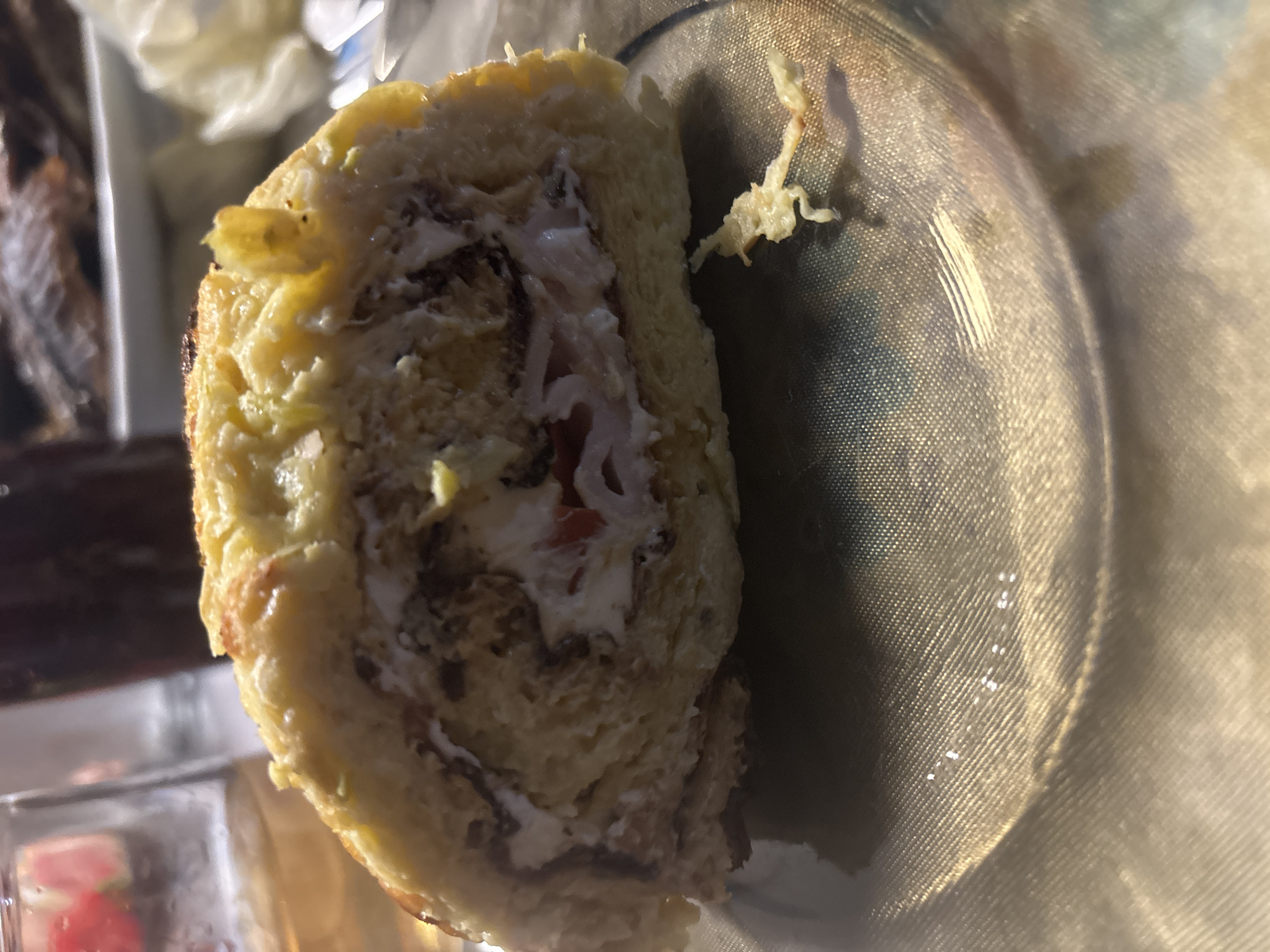 An overhead, medium close-up shot shows a slice of a roulade that appears to be a savory breakfast item. The roulade is cut on a diagonal and is the primary focus of the image. The outer layer is a golden-brown, slightly browned egg-based dough. Visible inside the roulade are layers of creamy white filling, interspersed with darker, possibly meat-based ingredients, and some red specks that could be tomato. The roulade rests on a textured, translucent gold-colored plate. To the left of the roulade, out of focus, is a glass containing a light-colored liquid and a red strawberry-like fruit. Behind the roulade, also out of focus, is a dark liquid in a bottle and a platter with what appears to be cooked fish. The background suggests a dining setting. The image does not provide any information about the city of Trudovets, Bulgaria.