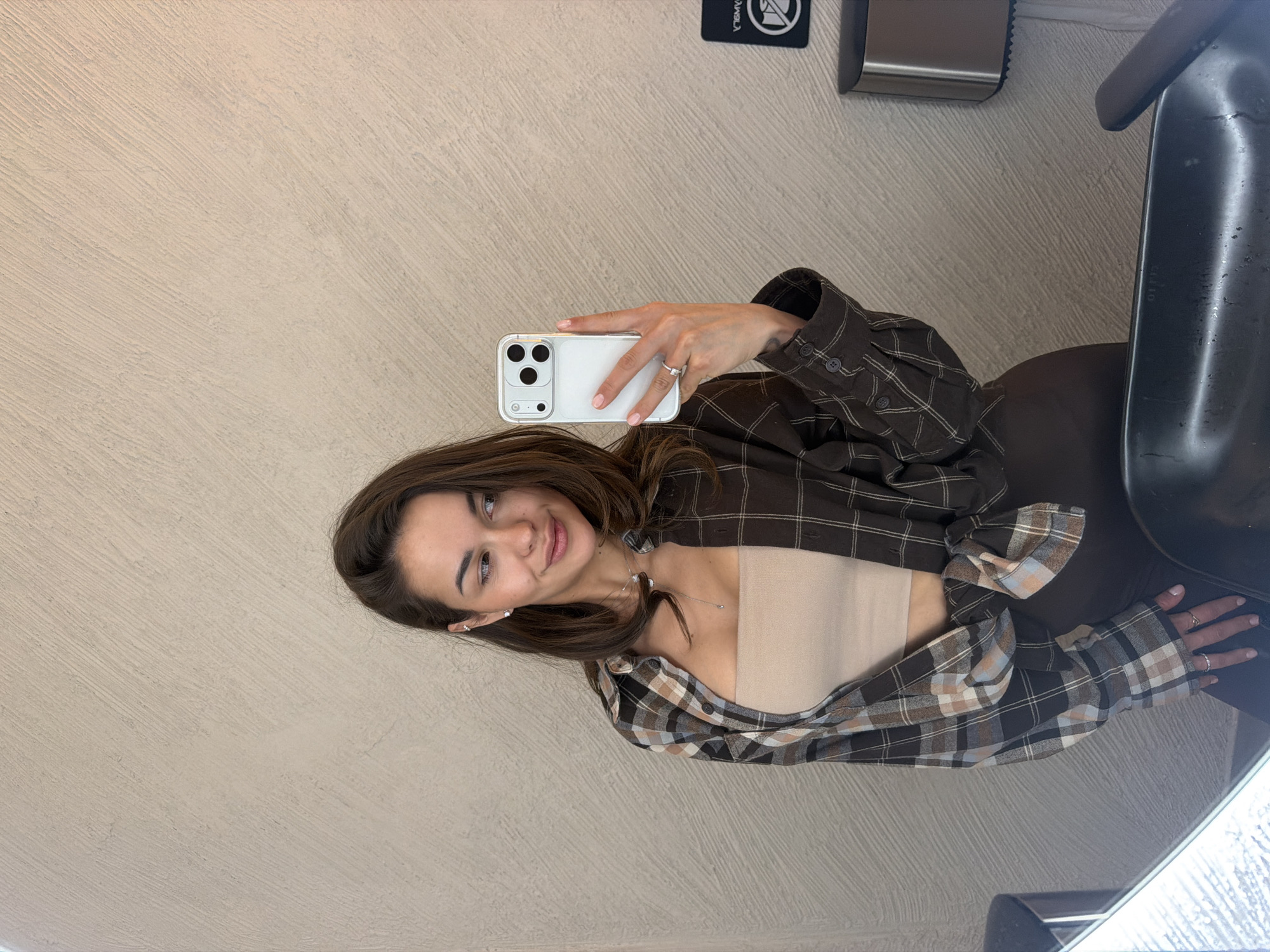 A young woman with long, dark brown hair is depicted in a selfie, likely taken in a mirror or reflective surface, showing her from the waist up. She has fair skin, a subtle smile, and is looking directly at her phone's camera. She wears a plaid collared shirt with a pattern of brown, black, light blue, and beige checks, unbuttoned over a light beige strapless top. Her accessories include small silver stud earrings, a delicate silver necklace with a small pendant, and a silver ring on her left ring finger. She holds a light-colored smartphone, featuring a distinct triple-camera setup, in her right hand.
The background reveals a textured light beige wall. In the upper right corner of the image (which would be the upper left if the image were upright), a black sign displays an inverted white "NO SMOKING" symbol and partially visible, inverted text, possibly "SANGHA". Below this sign, a metallic dispenser is mounted on the wall. To the woman's right, a dark, possibly black or dark brown, object resembling the back of a chair or seat is visible. The scene is an indoor setting in Krasnoyarsk, Russia.