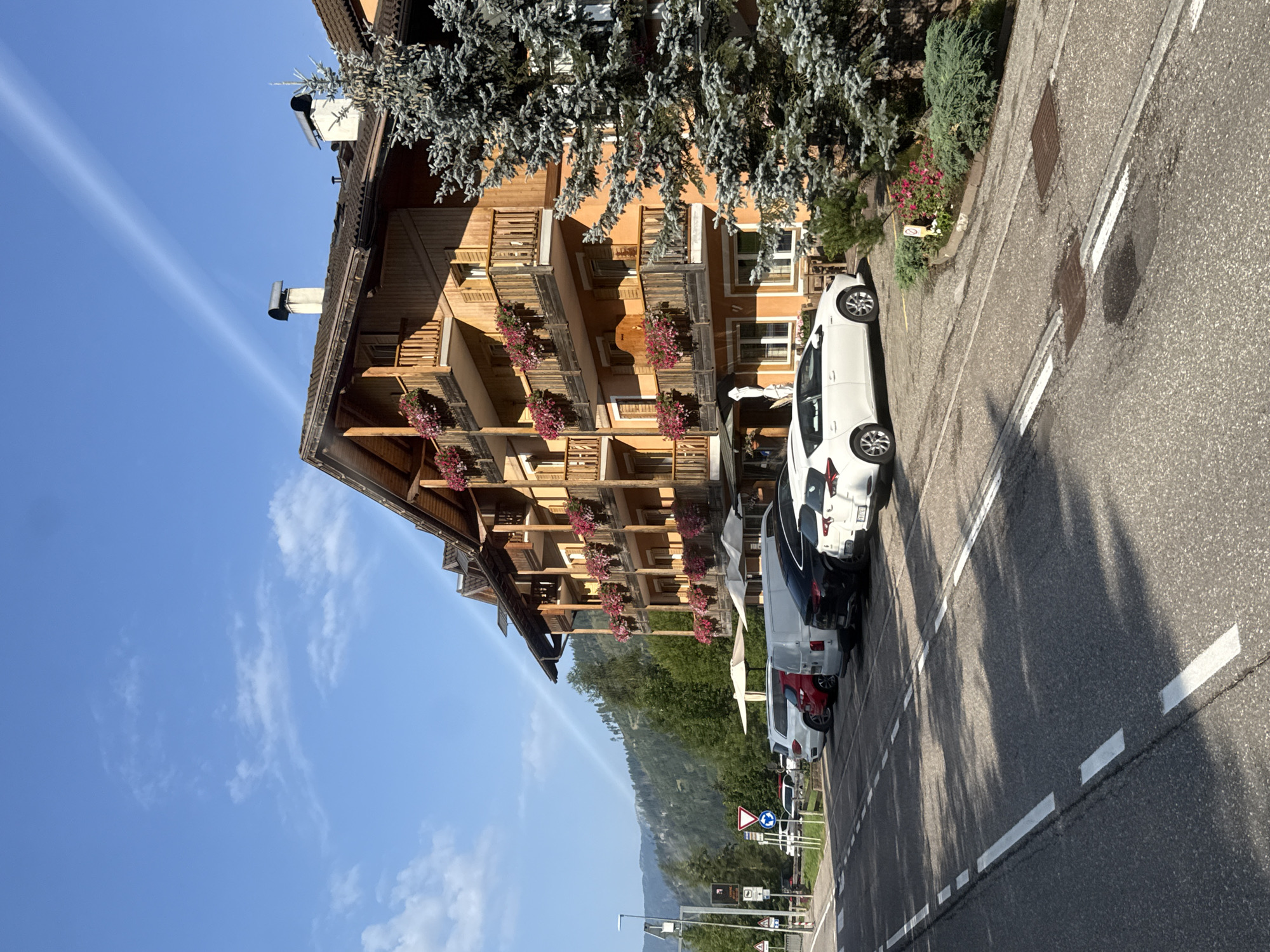 The image depicts a building with a wooden facade and balconies adorned with pink flowers, situated in Moena, Italy. In front of the building, a row of parked cars, including a white hatchback, a red SUV, a white van, and a black sedan, are visible. Adjacent to the building is a large evergreen tree. The scene also includes a paved road with white lane markings and a sidewalk. In the background, a lush green mountainside under a clear blue sky with scattered clouds completes the picturesque setting.