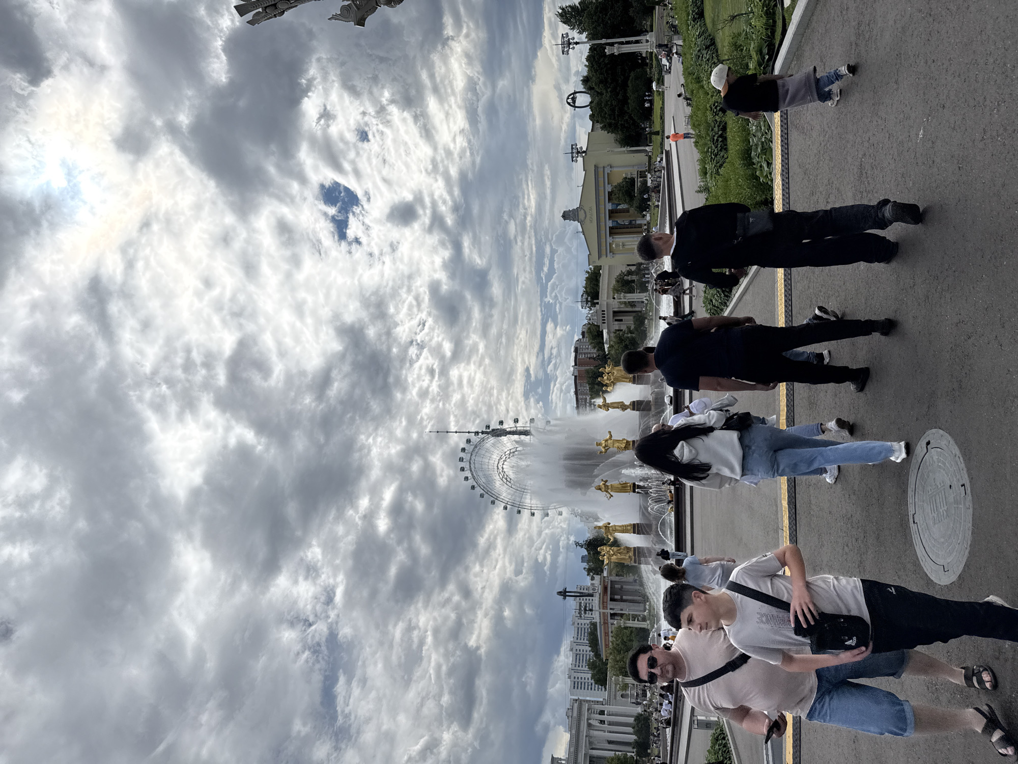 The image depicts a group of people in an outdoor setting with a large Ferris wheel and a fountain in the background. The sky is filled with dramatic, cloudy formations, suggesting it might be an overcast day with potential for rain. The perspective is from ground level, looking up towards the structures and people.
In the foreground, several individuals are visible. Two men are walking away from the camera, one wearing a dark jacket and the other a light-colored t-shirt. Another group, including a woman in a light-colored hoodie and jeans, is also present. Further back, a man in a black jacket is walking. There are many other figures scattered throughout the scene, indicating a public gathering or tourist area.
The prominent Ferris wheel is a significant landmark, suggesting the location is likely an amusement park or a large public space. The fountain with golden statues adds to the grandeur of the scene. Buildings with architectural details can be seen behind the fountain, some appearing to be of classical design. The overall impression is a busy, lively atmosphere, possibly during a warm day given the attire of some individuals. The orientation of the image suggests it may have been taken with the camera held vertically. No discernible text is visible in the image. The location context is Moscow, Russia.