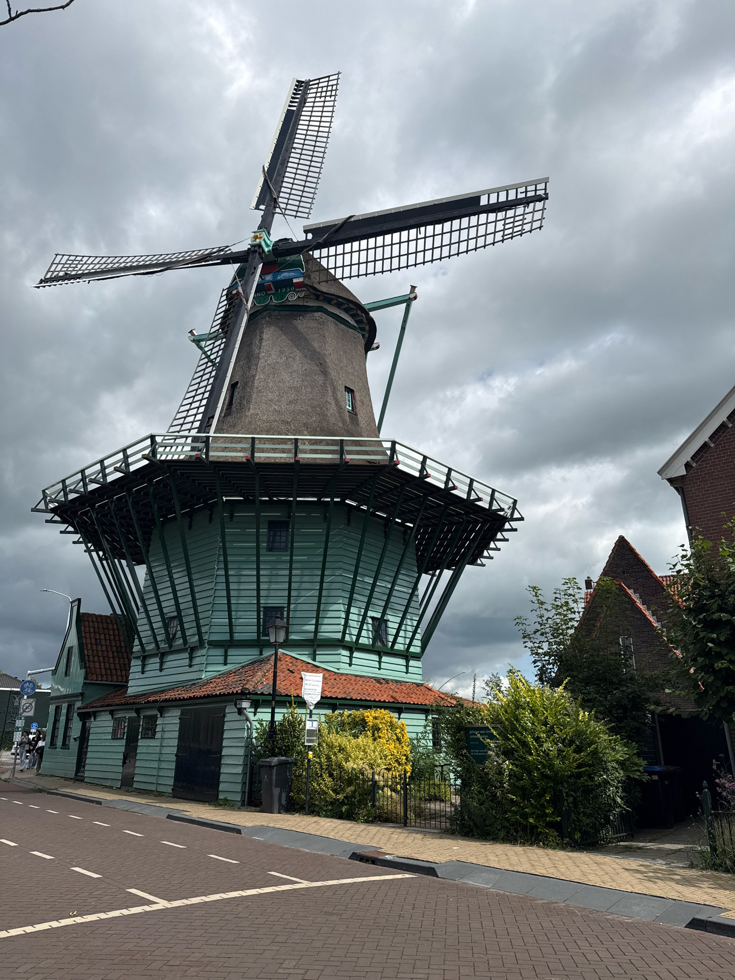 The image shows the Molenaars, a Dutch windmill located in Zaandijk, Netherlands. The windmill features a thatched roof and wooden sails, with a green painted base. Adjacent to the windmill is a traditional Dutch house with a red tiled roof. The scene is set against a cloudy sky. A street with brick paving is visible in the foreground, with a few people walking on the sidewalk. The overall impression is a picturesque historical Dutch streetscape.