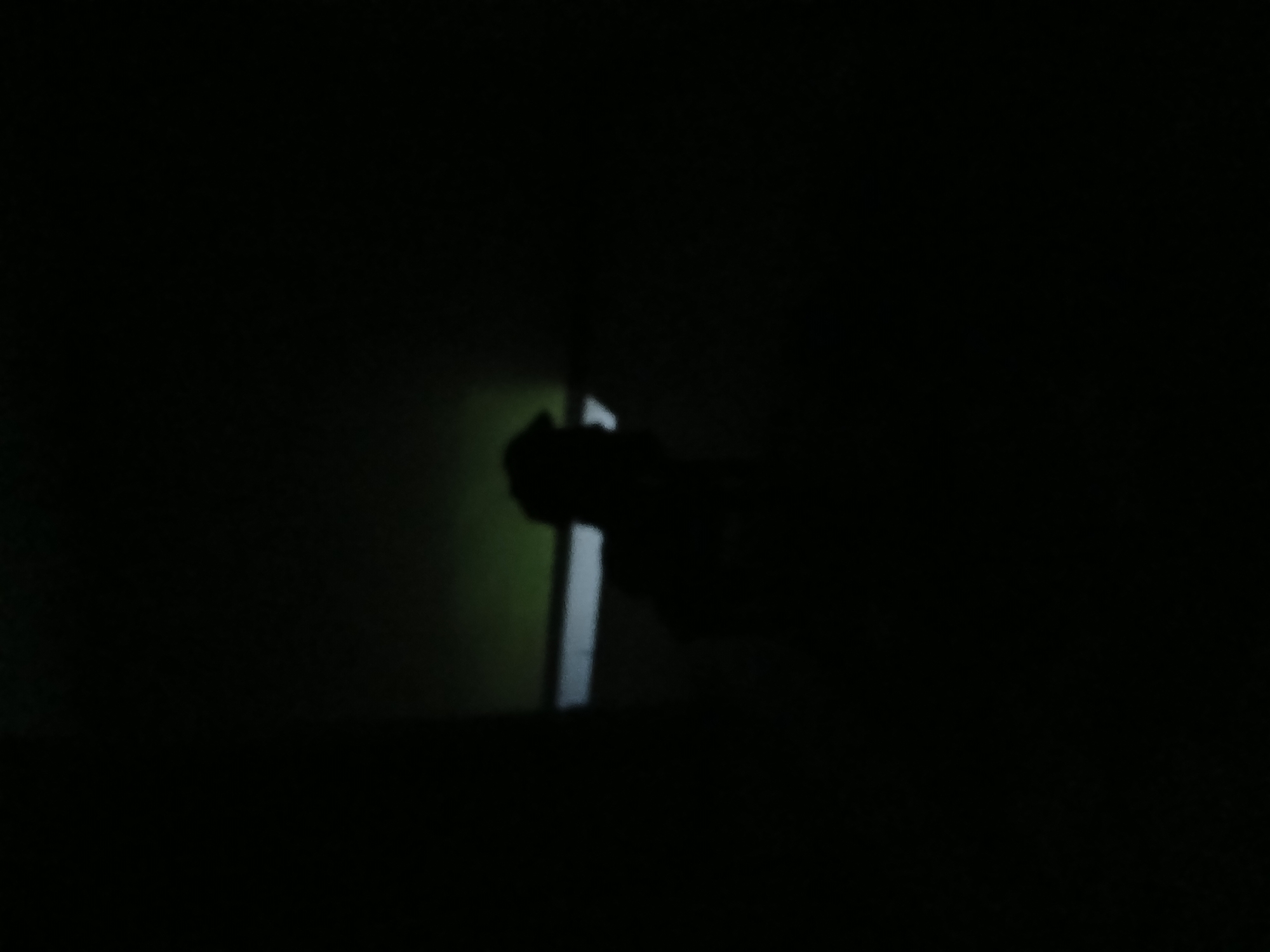 The image depicts a dark, dimly lit environment. A silhouette of a person, likely wearing a cap, is visible in the center of the frame. They appear to be in a seated or hunched position. To the left of the silhouette, a horizontal line of light or a reflective surface is present. The background is predominantly dark, with a faint greenish glow in the upper portion of the frame, suggesting an artificial light source or ambient light filtering through. The overall impression is that of a clandestine or low-light setting.