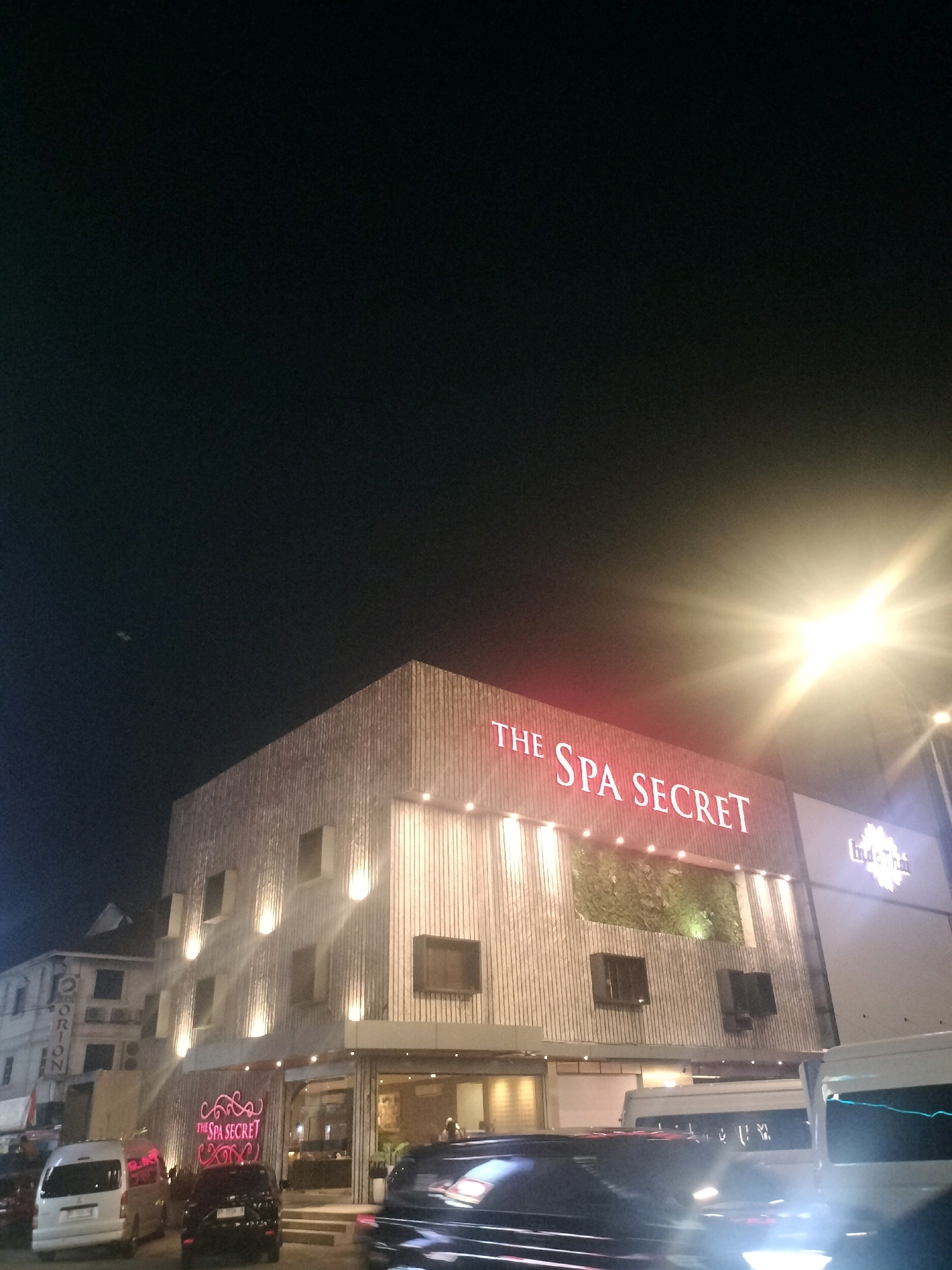 The image depicts a nighttime scene in Batam, Indonesia, focusing on the facade of "THE SPA SECRET," a commercial establishment. The building features a modern architectural design with a textured wooden exterior. Prominently displayed on the upper facade is the neon sign "THE SPA SECRET" in red lettering. Below this, a green, living wall or moss panel adds a natural element to the building's design. Several windows are visible on the upper floors, and accent lighting illuminates the building's facade.
In the foreground, several vehicles are present, including white vans and a black SUV, suggesting this is a street-level view. The vehicles are partially illuminated by streetlights and the bright glare of a high-intensity light source, possibly a street lamp or vehicle headlight, which creates a lens flare in the upper right portion of the image. Adjacent to "THE SPA SECRET," a sign for "IndoThai" is partially visible, indicating a neighboring business. The background shows other buildings, including a structure with the word "ORION" visible, suggesting a commercial or mixed-use area within the city. The sky is dark, consistent with a nighttime setting.