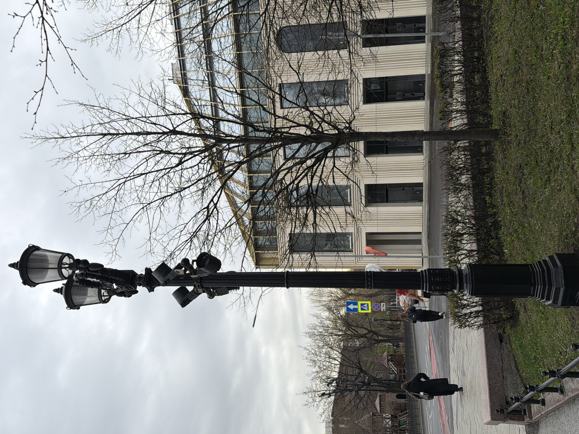 This image captures an urban street scene in Moscow, Russia, under an overcast sky, likely during late winter or early spring due to the bare trees. The foreground features a tall, ornate black streetlamp with multiple decorative light fixtures. Its dark silhouette stands against the bright, cloudy sky.
Intricate bare branches of several deciduous trees dominate the midground and upper sections of the image, partially obscuring a modern multi-story building behind them. This building has a facade of light-colored horizontal panels and large, dark-framed windows. To its left, another section of the building displays a curved, reflective glass exterior.
Running parallel to the building is a strip of green grass or low hedges, followed by a paved sidewalk. Two individuals, dressed in dark clothing, are visible walking on this sidewalk, engaged in routine activity. Beyond the sidewalk lies a road with a painted pedestrian crossing. Road signs are also visible; one is a blue square sign with a white left-pointing arrow and a pedestrian crossing symbol, indicating a directional pedestrian crossing. Another standard road sign, a red circle with a blue center and a red diagonal line, signifies "No Stopping." The overall impression is one of a typical urban environment during a cool, possibly damp, overcast day. No other specific text is legible.