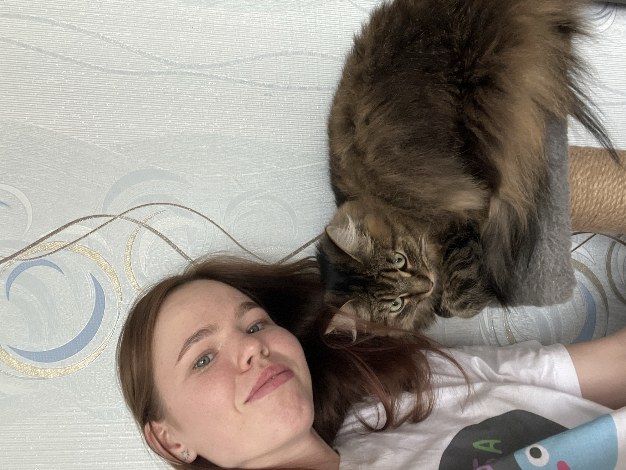 A woman and a cat are lying down. The woman is on her back, looking up and smiling at the camera. She has reddish-brown hair and is wearing a white t-shirt with a cartoon design on it. The cat, a large, brown tabby, is perched above her, also looking at the camera. The background is a wall with a patterned wallpaper. Based on other information, it's likely this photo was taken in Chaikovsky, Russia.