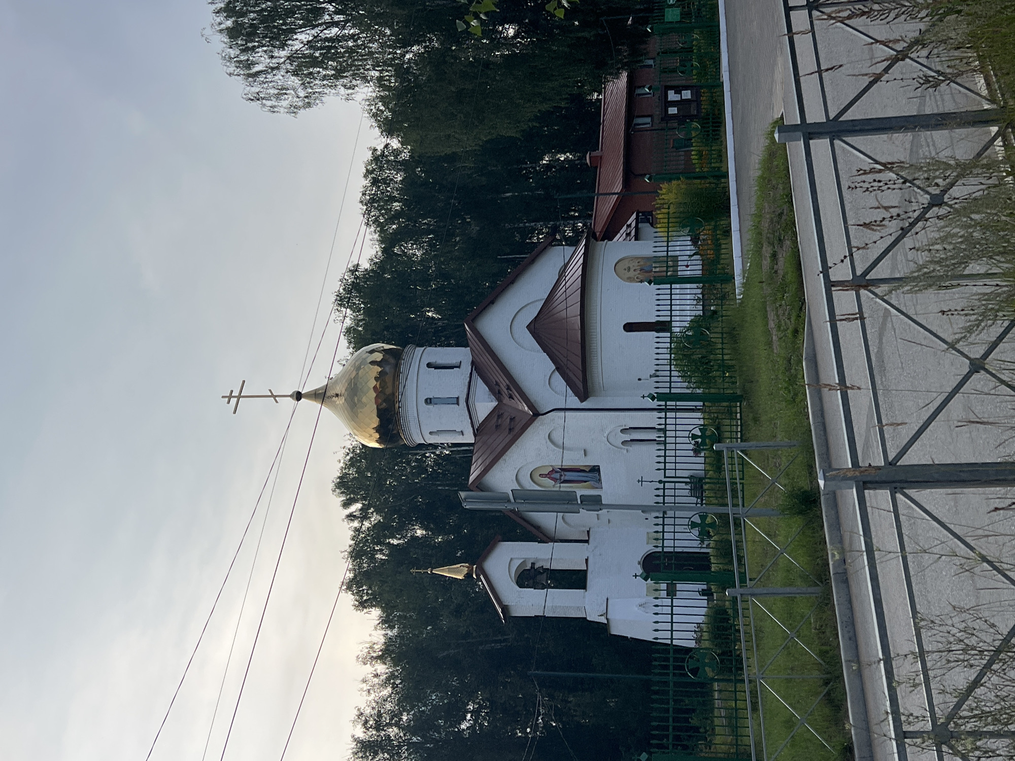 The image shows a white Orthodox church with a golden dome and a copper-colored roof, located in the city of Noviy, Russia. The church features a bell tower on the left and a smaller domed structure on the right. Religious icons are visible on the exterior walls. The church is enclosed by a green metal fence. Behind the church, a dense line of green trees forms the background. A red-brick building is partially visible to the right of the church. In the foreground, a street and a gray metal fence are present. Overhead power lines are visible against the overcast sky.