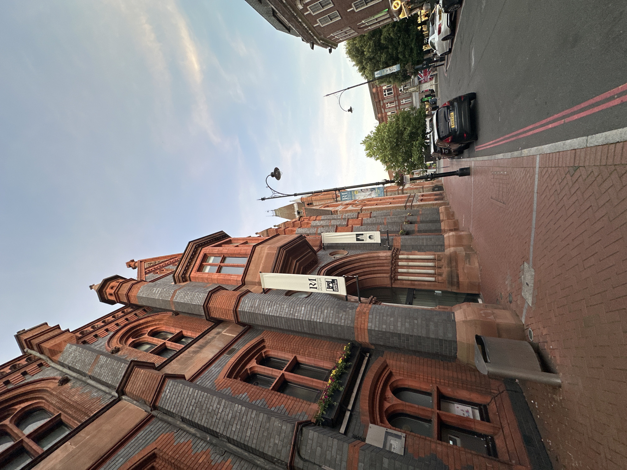 A low-angle, wide shot captures the facade of a red brick and grey stone building on the left, adorned with arched windows and decorative architectural elements. Several white banners with black text hang from the building. A brick sidewalk runs along the building, with a modern silver trash receptacle positioned near the corner. The sidewalk transitions to a street on the right, where two cars are parked and a white car and a dark SUV are driving. Lampposts line the street, and trees and other buildings are visible in the background. The sky above is a pale blue with wispy clouds.