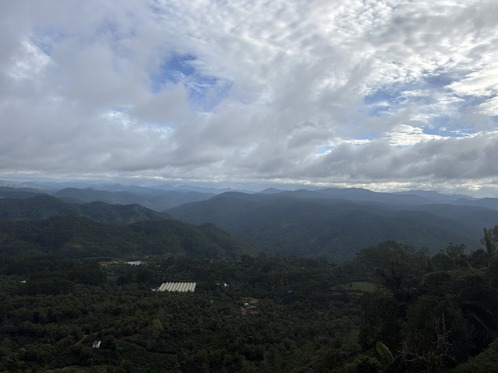 This is a panoramic, high-angle, outdoor photograph taken on a cloudy day, looking out over a vast expanse of rolling, densely forested mountains. The foreground is dominated by dark green trees, with a small white building visible in the lower left quadrant, appearing to be a solitary house or structure. Further into the middle ground, there are structures that appear to be greenhouses or agricultural buildings with white, translucent coverings, indicative of farming or cultivation. The landscape recedes into the distance with multiple layers of mountains fading into a hazy blue-gray. The sky is filled with a dramatic mix of white and gray clouds, with patches of blue sky peeking through. The scene is consistent with the mountainous terrain found in the region of Phát Chi, Vietnam, known for its agricultural landscapes and natural beauty. There are no people or animals visible in the image.