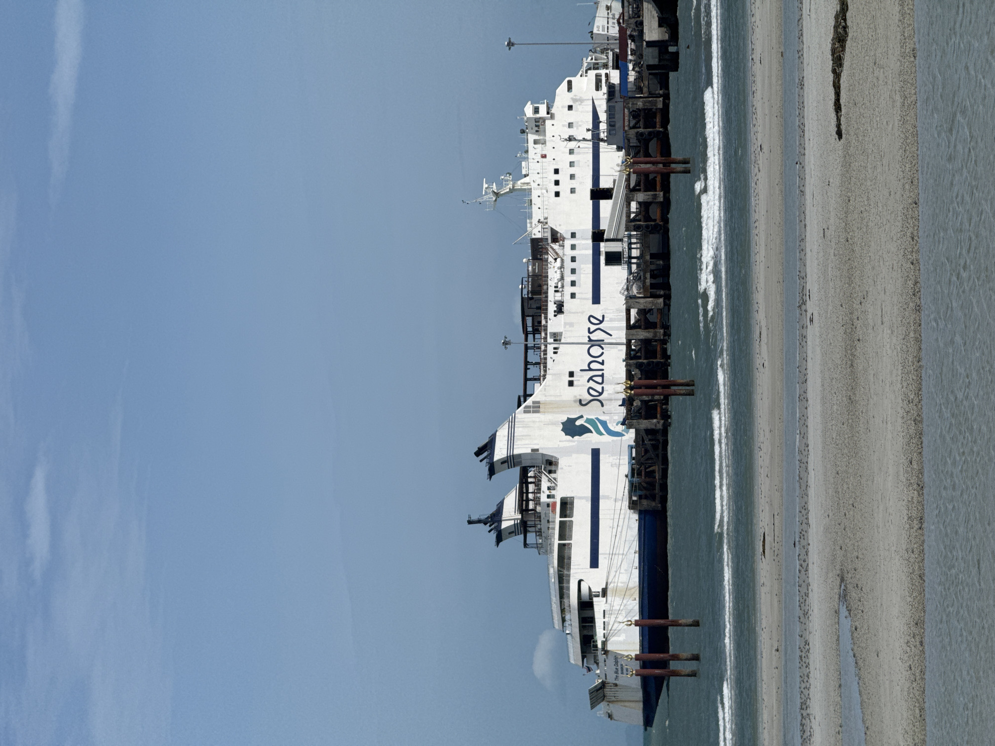 A large white ferry named "Seahorse" is docked at a pier on the coast of Ko Samui District, Thailand. The ferry has a blue stripe running along its side and a logo of a seahorse. The pier is made of wood and concrete, with several vertical posts in the water. The water is choppy, with small waves. The sky is clear and blue with a few wispy clouds. In the foreground, there is a sandy beach with some water. The scene appears to be a sunny day.