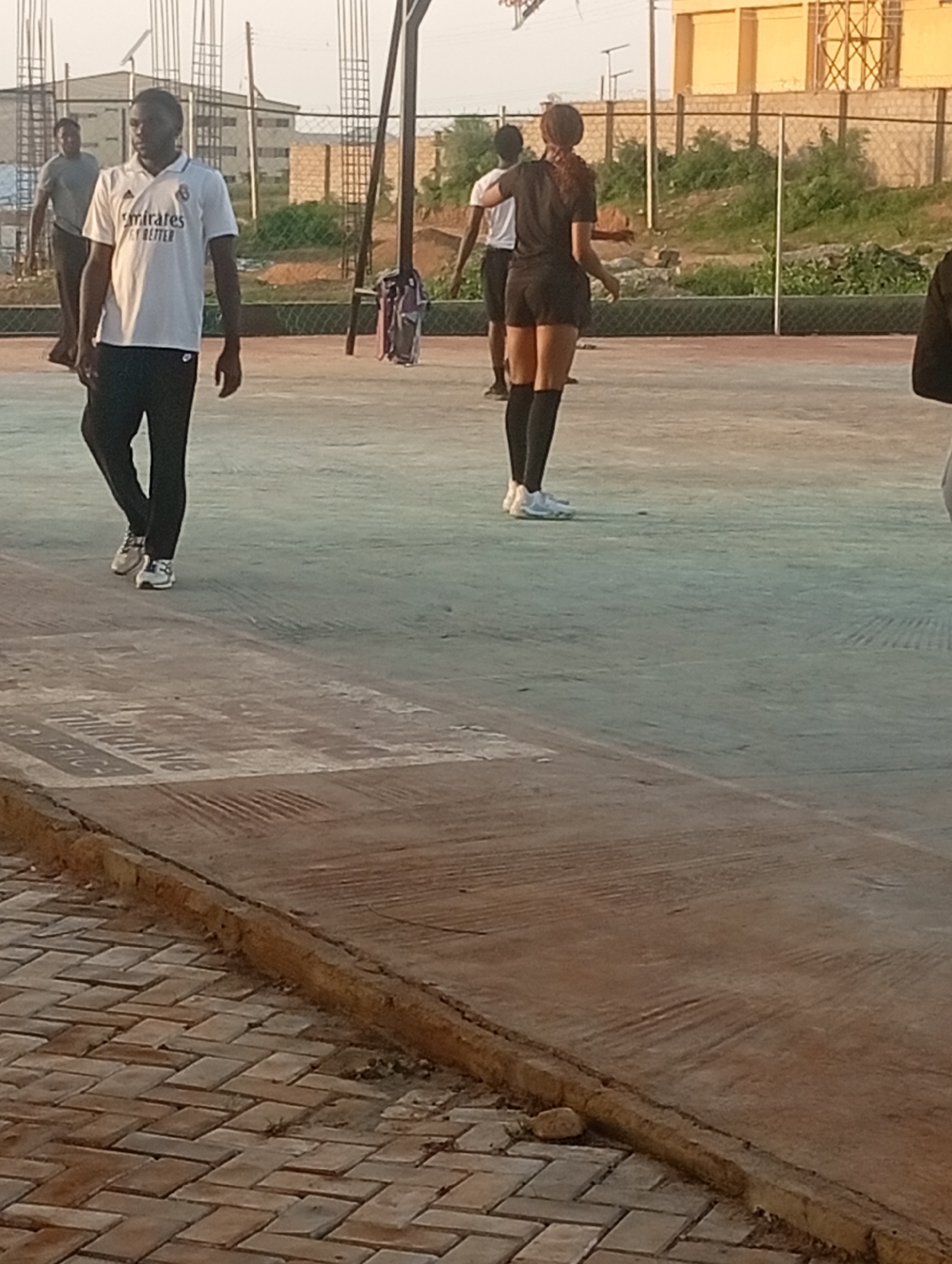 A medium shot shows four individuals on a hard surface, possibly a tennis or basketball court, with a brick pathway in the foreground and a chain-link fence in the background. The lighting suggests it is late afternoon or early morning.
In the foreground, a man wearing a white t-shirt with "Emirates Fly Better" emblazoned on it and black pants walks to the left. He has dark hair and appears to be in his early twenties. To his left and slightly behind him, another man in a grey t-shirt walks away from the camera.
Further back, a woman in a black t-shirt and black shorts with knee-high black socks and white sneakers is facing away from the camera, appearing to engage in some activity. Beside her, a person wearing a black top and black pants is partially visible on the far right of the frame.
Behind the individuals, a chain-link fence separates the court from a grassy area with some buildings and construction structures in the distance. The overall setting is an outdoor urban environment.