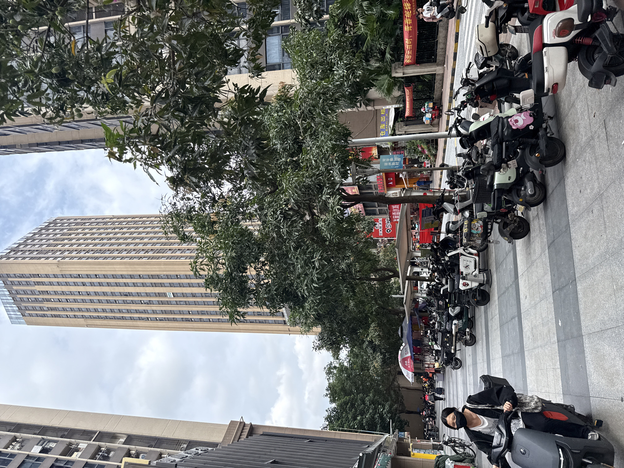 In Haizhu District, China, a woman wearing a black mask and black jacket rides a dark gray electric scooter on a gray and white checkered pedestrian walkway. She is carrying a patterned tote bag over her left shoulder. Behind her, a row of parked electric scooters and bicycles lines the walkway. In the background, a tall, tan skyscraper with numerous windows dominates the skyline, flanked by other tall buildings and lush green trees. A street with a yellow and black striped pedestrian crossing is visible beyond the parked vehicles, with a few people walking and a red and white striped umbrella over a vendor stall. The sky is overcast with clouds.