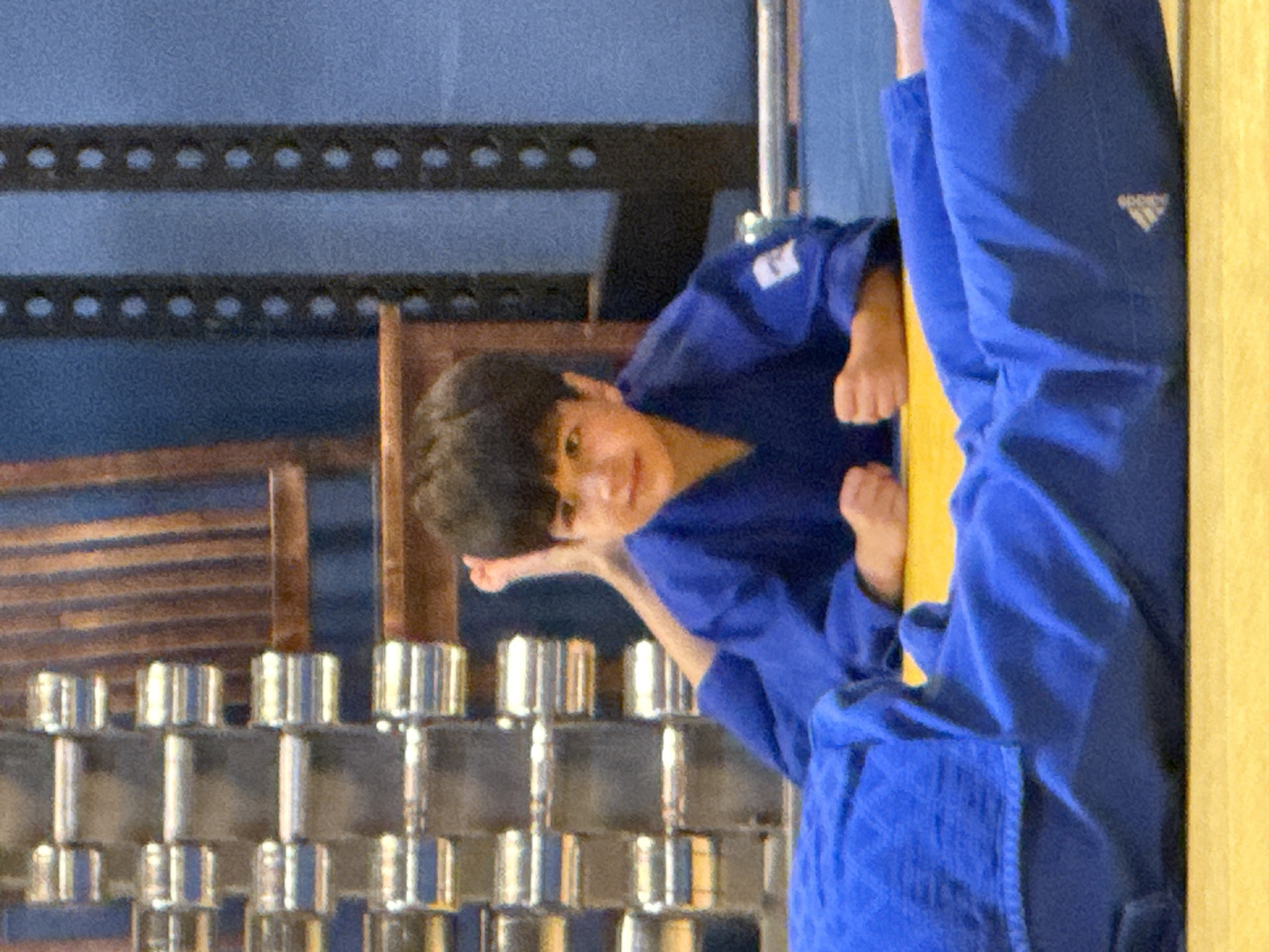 A young boy wearing a blue judogi lies on his back on a yellow mat. His head is turned to his right, and he is looking at the camera with a slight smile. His right leg is bent at the knee, and his foot is visible. His left arm is bent at the elbow, and his hand is resting on his chest. His right arm is bent at the elbow, and his hand is resting on his thigh. In the background, there are rows of weights and a wooden bench. The boy appears to be in a gym or dojo.