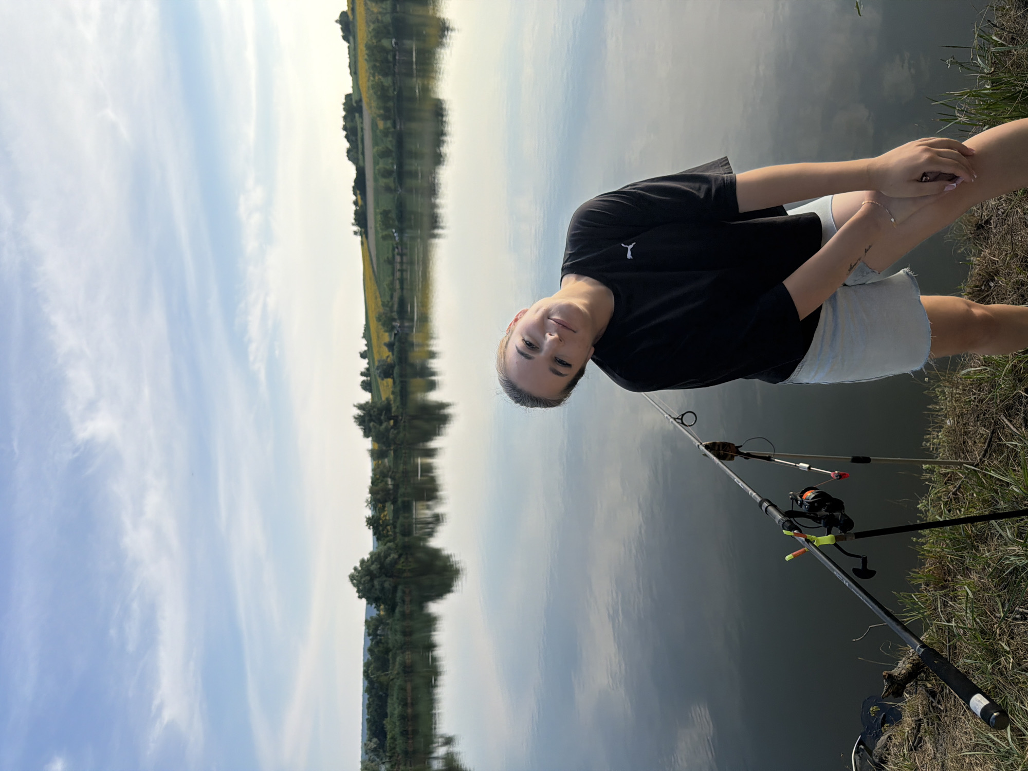 A young woman with blonde hair pulled back poses by a lake with fishing rods in the foreground. She is wearing a black t-shirt and light blue denim shorts. The lake reflects the sky and the distant treeline. The background features rolling hills with fields of sunflowers under a partly cloudy sky. The overall scene suggests a relaxed outdoor setting, likely a fishing spot in Tătărăști, Moldova.