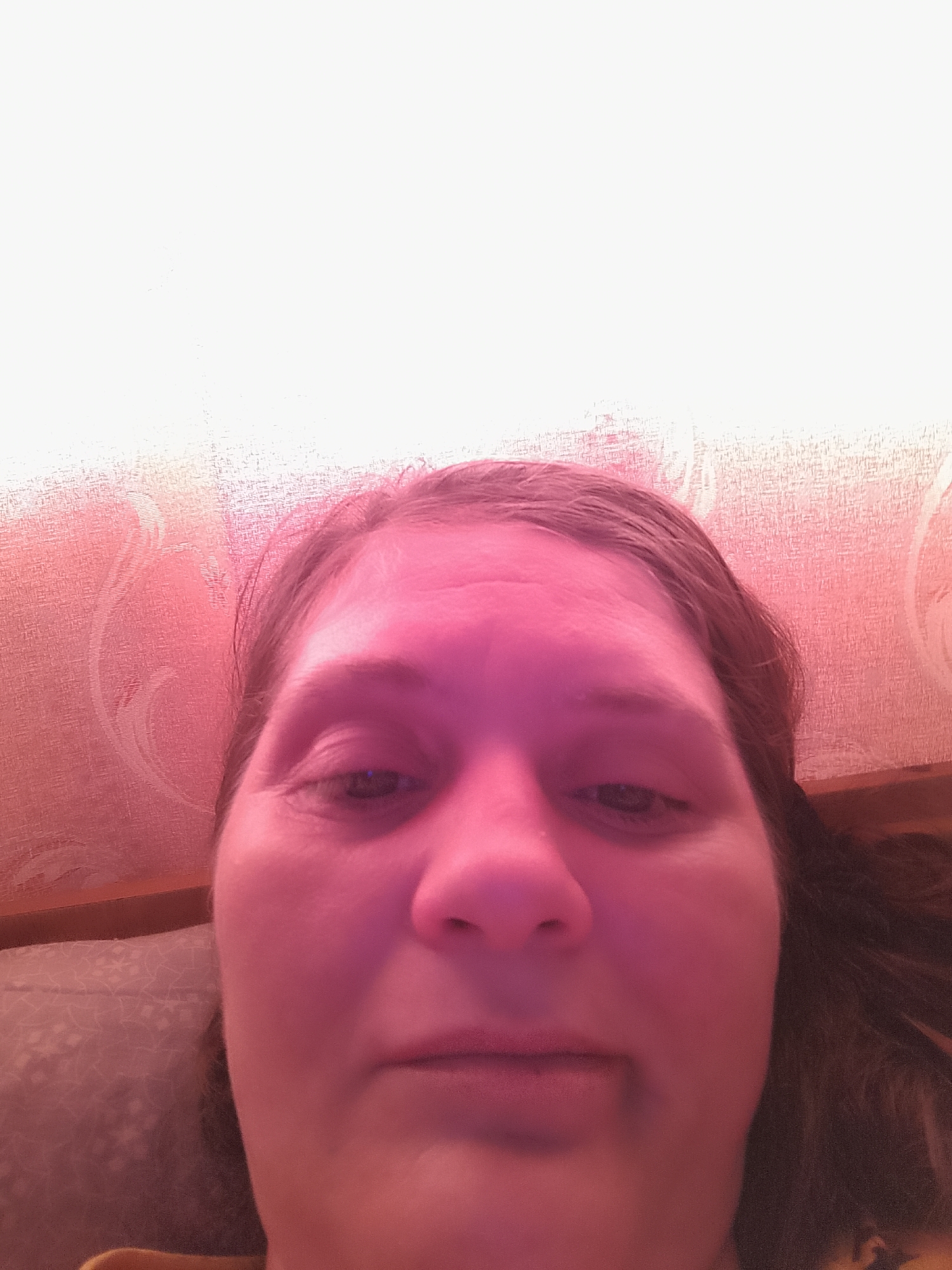 A close-up, low-angle, poorly lit selfie shows the face of a woman. The woman has long brown hair, is looking slightly to the left, and has her mouth slightly ajar. She is wearing a yellow top. The background appears to be a wall with a floral patterned wallpaper, lit with a pink light.