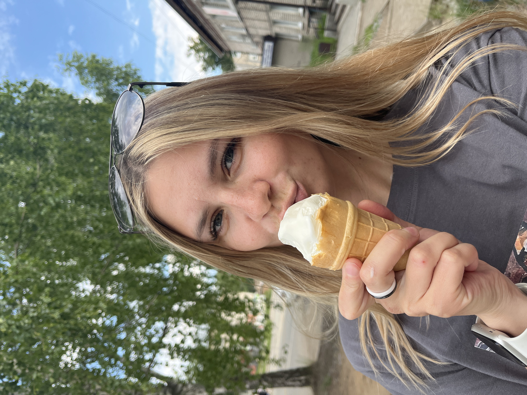A young woman with blonde hair is holding an ice cream cone to her mouth. She is wearing a grey t-shirt and a ring on her left hand. Sunglasses are perched on top of her head. She appears to be outdoors, with trees and buildings in the background. The scene is likely a street in the city of Chaikovsky, Russia, given the style of the building and the presence of birch trees common in the region. The sky is blue with some clouds, suggesting it is daytime.
