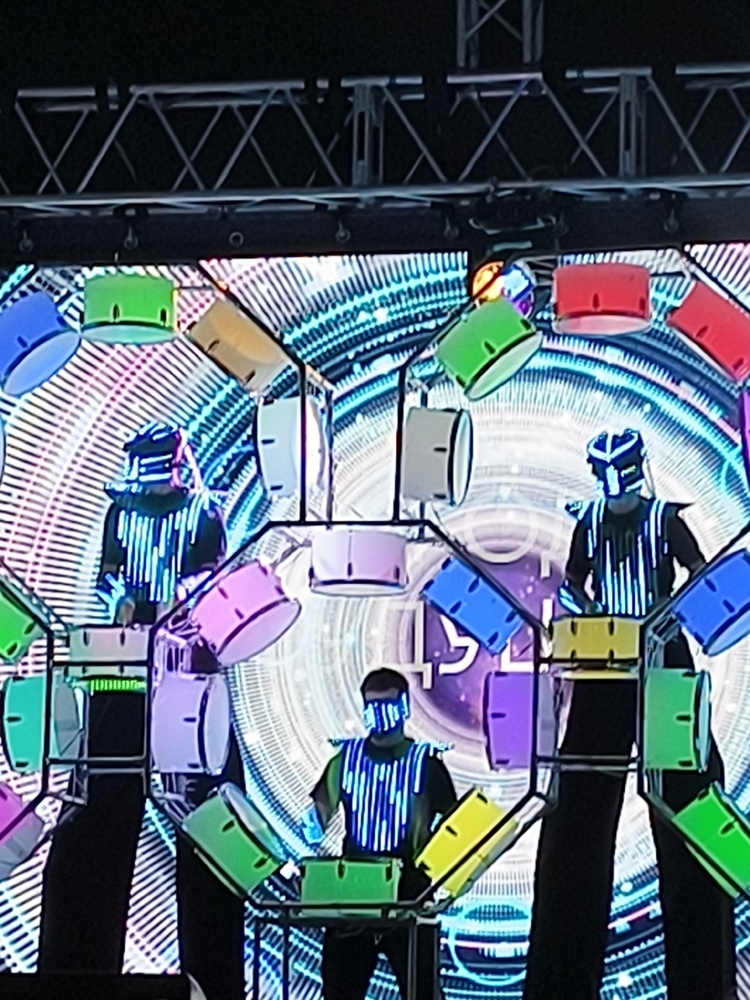 The image captures a performance art or concert scene where several individuals are dressed in futuristic, illuminated costumes, appearing to be playing electronic drums. The stage is set against a large, vibrant, digital backdrop with geometric patterns and bright blue and white lights, creating a high-tech and energetic atmosphere.
In the foreground, two performers are centrally positioned. The one on the right is wearing a black outfit adorned with vertical blue LED stripes, a matching helmet with a visor, and shoulder pads. They are standing behind a setup of yellow and green electronic drum pads, seemingly in the act of playing them. The performer on the left, also in a similarly styled costume, is partially visible and appears to be engaged in the performance as well.
Surrounding the performers, and suspended above them, are various colorful objects that resemble drum shells. These include green, blue, red, yellow, and pink cylindrical objects, some of which also appear to have light elements. These are arranged in a dynamic, almost chaotic, yet organized manner, contributing to the overall visual spectacle.
The backdrop is a complex array of illuminated lines and circular patterns, giving the impression of a digital or futuristic interface. The lighting is predominantly cool-toned, with blues and whites dominating, interspersed with the bright colors of the drum elements and costumes, creating a strong contrast and visual depth. The overall impression is one of a modern, technologically advanced performance focused on electronic music and visual arts. The location is not specified beyond "Unknown, Unknown".