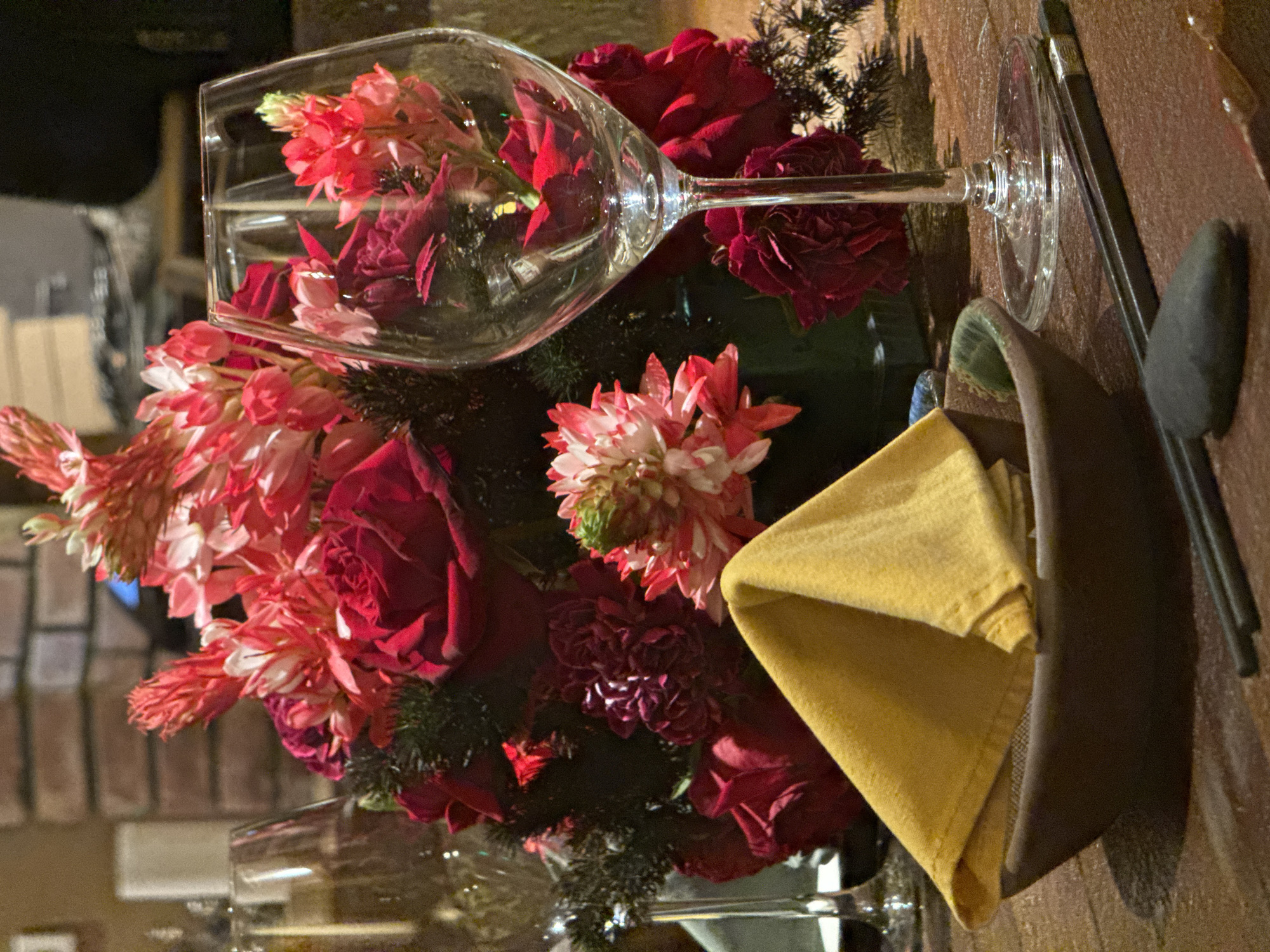 A still life arrangement on a dark wood table features a wine glass lying horizontally, partially filled with pink and red flowers. A bouquet of red roses and pink flowers surrounds the glass. Next to the arrangement is a dark brown napkin holder with a folded yellow napkin. Cutlery, including a spoon and knife, lies beside the napkin holder. The image is taken from a close-up perspective, with ambient lighting illuminating the scene. Brickwork can be seen in the background suggesting this is inside a building in Hà Nội, Vietnam.