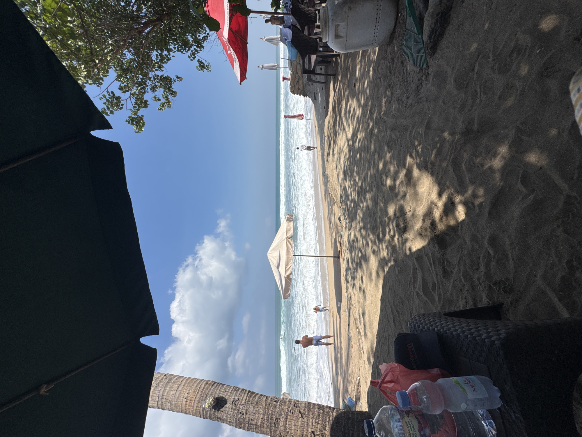A medium shot captures a beach scene in Kuta, Indonesia, under a bright blue sky with scattered clouds. In the foreground, a dark umbrella obscures the top of the frame, casting shadows on the sandy ground. To the left, a tall palm tree trunk is visible. On a dark woven table in the immediate foreground, two plastic water bottles and a red plastic bag are present.
Further back, a light-colored umbrella stands on the sand, with a man in blue shorts and a woman in a blue swimsuit standing near the water's edge, facing the ocean. A child in a light blue outfit is also near the water. The ocean exhibits moderate waves, and the horizon is a clear demarcation between the sea and sky.
To the right, several beach chairs and tables are set up under a large red umbrella, with more white umbrellas in the background. Sandbags are piled up behind the seating area. A large white container and a green rake are also visible on the sand. Another person is standing near the seating area, looking out at the sea. The scene suggests a relaxed atmosphere at a beachside establishment.