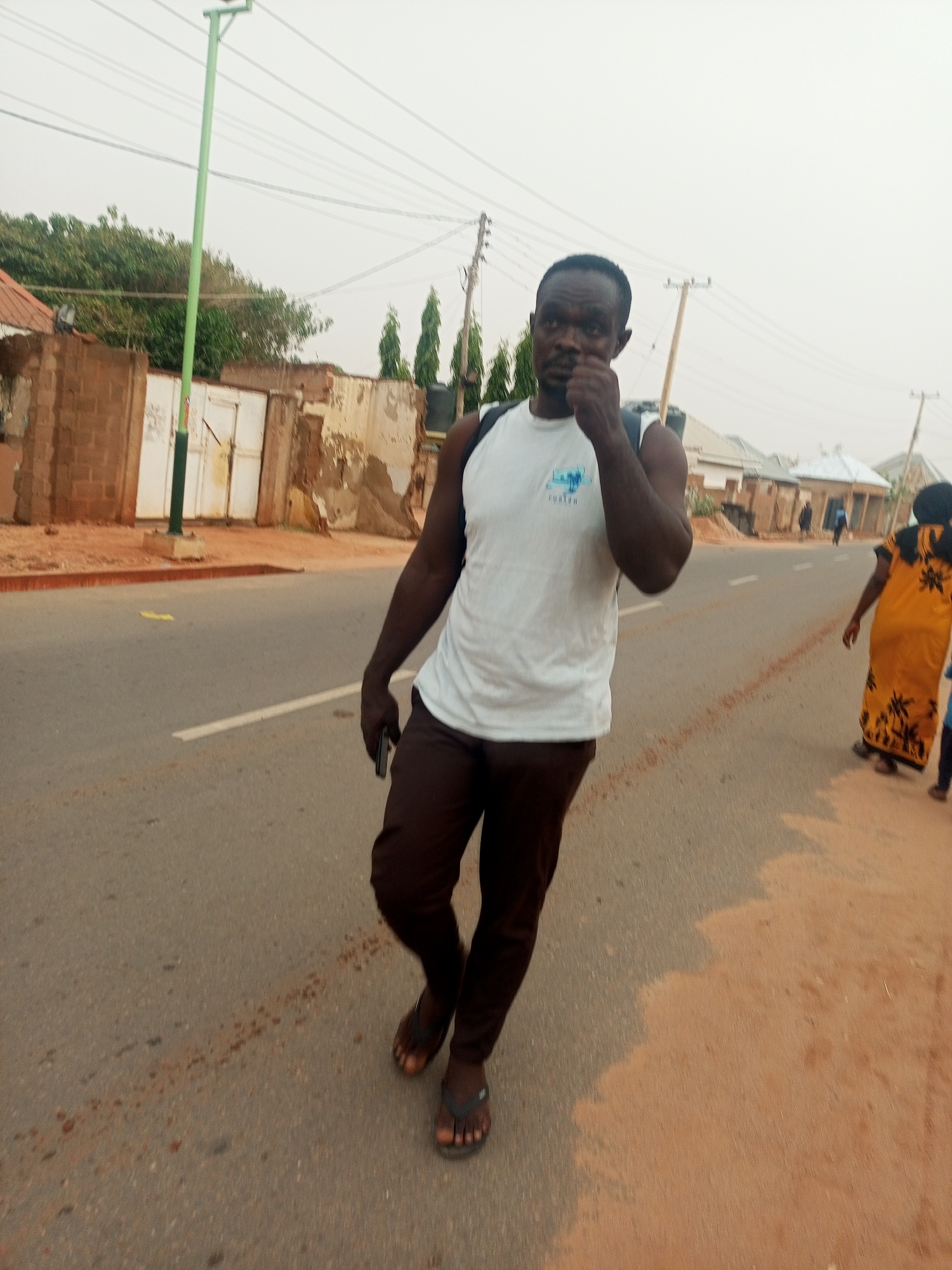 A man is walking on a street in what appears to be a residential area. He is wearing a white sleeveless shirt, brown pants, and sandals. He has a backpack on his back and is holding a cell phone in his left hand. He is walking on the left side of the road, facing the camera, with a slightly blurred background showing buildings, power lines, and other people. The environment seems to be an urban or suburban area with dusty roads and a faded sky. The location is identified as Tempure, Nigeria.