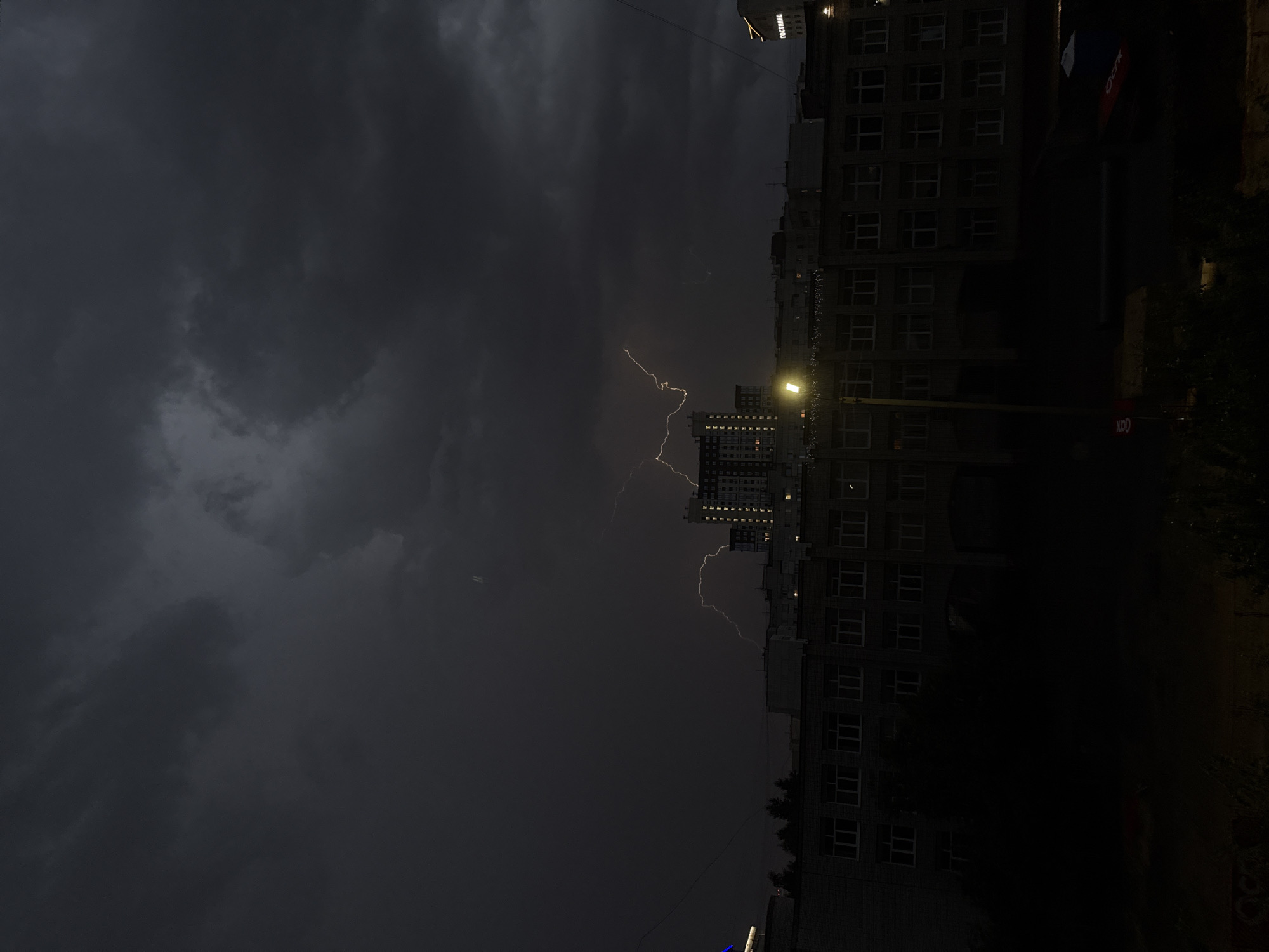 The image shows a dramatic nighttime cityscape with a fierce thunderstorm. Jagged bolts of lightning illuminate the dark, cloudy sky above a collection of buildings. In the foreground, a multi-story brick building with numerous windows is visible, partially silhouetted against the storm. A streetlamp casts a warm glow near the center of the frame, its light reflecting on the wet surfaces. Further back, taller buildings with illuminated windows rise into the darkness, creating a stark contrast with the storm's power. The overall atmosphere is one of intense weather and urban activity under the cover of night.