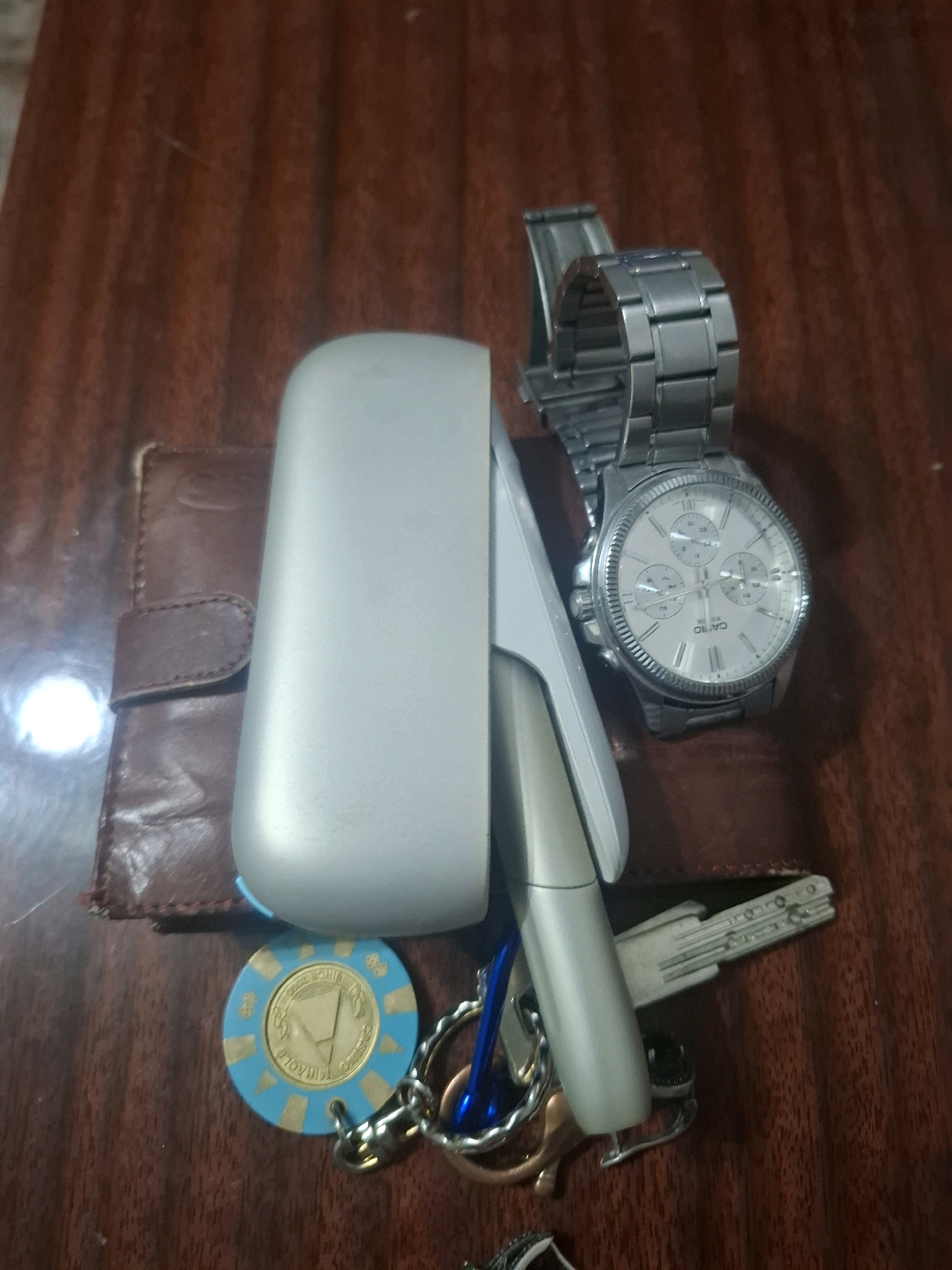 The image displays a collection of personal items on a wooden surface. A silver-toned watch with a metal band is prominently featured. Next to it is a light gray electronic device, possibly a vape or e-cigarette, and a brown leather wallet. A circular, blue and gold token, resembling a casino chip, is also visible, along with several keys attached to a keyring. One key is a car key with a silver casing and integrated buttons. The overall impression is that of everyday objects laid out on a table, likely in a domestic or personal setting. The mention of Almaty, Kazakhstan, is not supported by any visual evidence in the image itself.