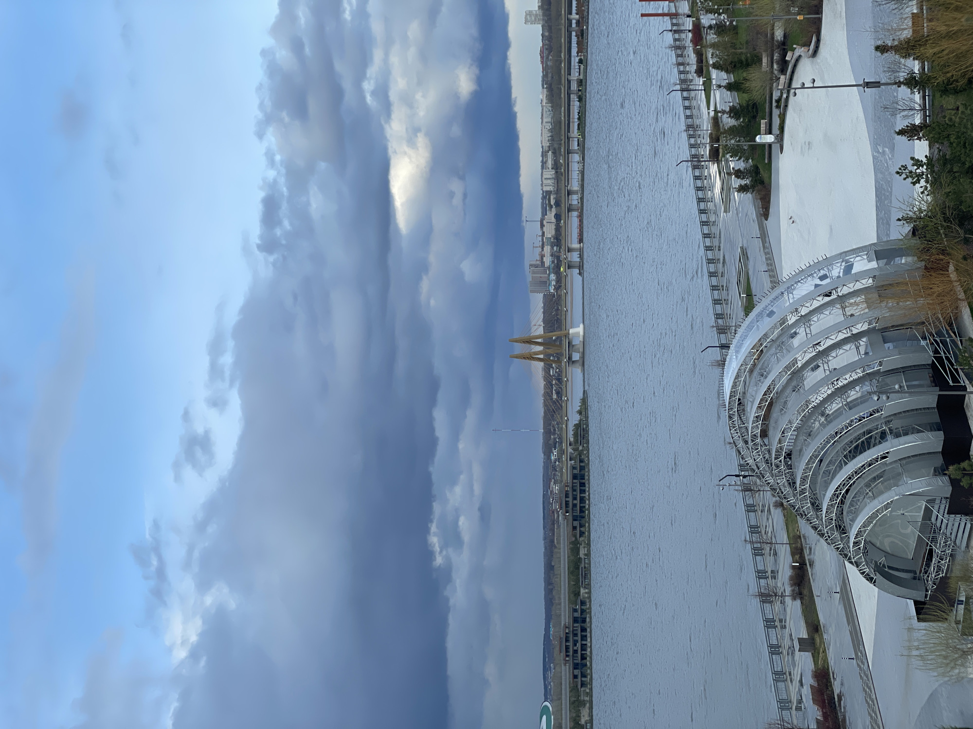 The image depicts an urban waterscape under an overcast sky. A wide body of water, likely a river or large lake, occupies the central and lower portions of the frame, its surface rippled by light breezes.
Across the water, a distant city skyline is visible, featuring numerous buildings and structures, including a prominent bridge with distinctive yellow or golden, arch-like supports. Along the near bank of the water, a modern, large-scale structure with a distinctive curved, ribbed design dominates the right side. This building appears to be made of white or silver-toned panels and framework, possibly indicating a stadium, cultural center, or public amenity. Adjacent to this structure, a paved promenade or pathway runs parallel to the water's edge, dotted with light posts, railings, and scattered trees and shrubs.
The sky above is dramatic, filled with heavy, dark grey clouds that stretch across much of the upper frame, contrasting with lighter patches of blue sky peeking through. The overall lighting suggests an overcast day, possibly late afternoon or an impending weather change. No specific human activity or visible text can be clearly identified. The scene portrays a developed urban environment set against a vast body of water, under a dynamic and cloudy sky.