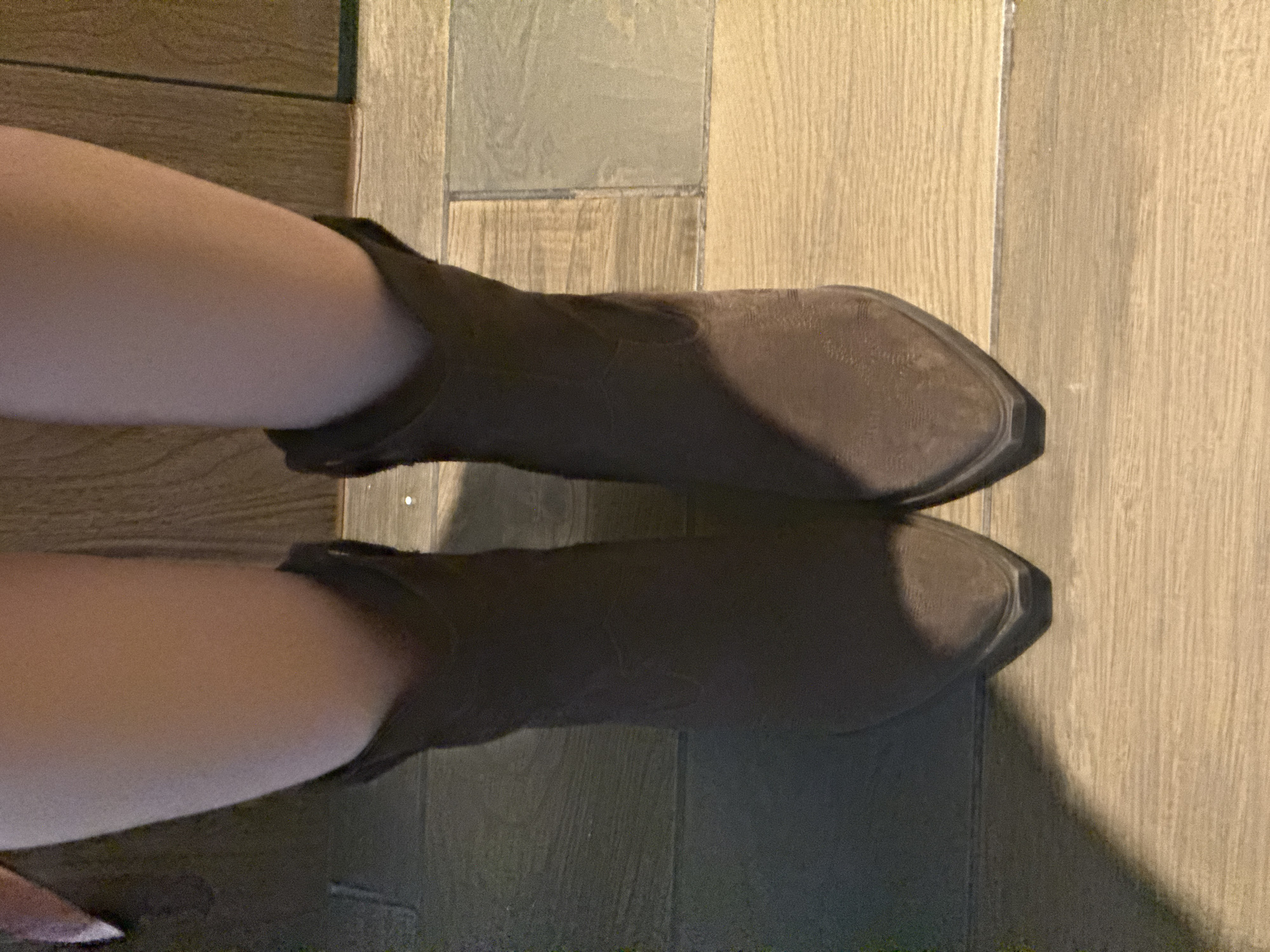 A low-angle, indoor shot depicts two legs wearing ankle boots, presumably on a person. The legs are positioned horizontally across the frame. The boots are a dark brown color with a pointed toe. The background is a wooden floor, likely made of planks or tiles with a light tan or beige tone. The lighting is soft and diffused. The location is unknown and there is no indication that the image was taken in Mexico City, Mexico.