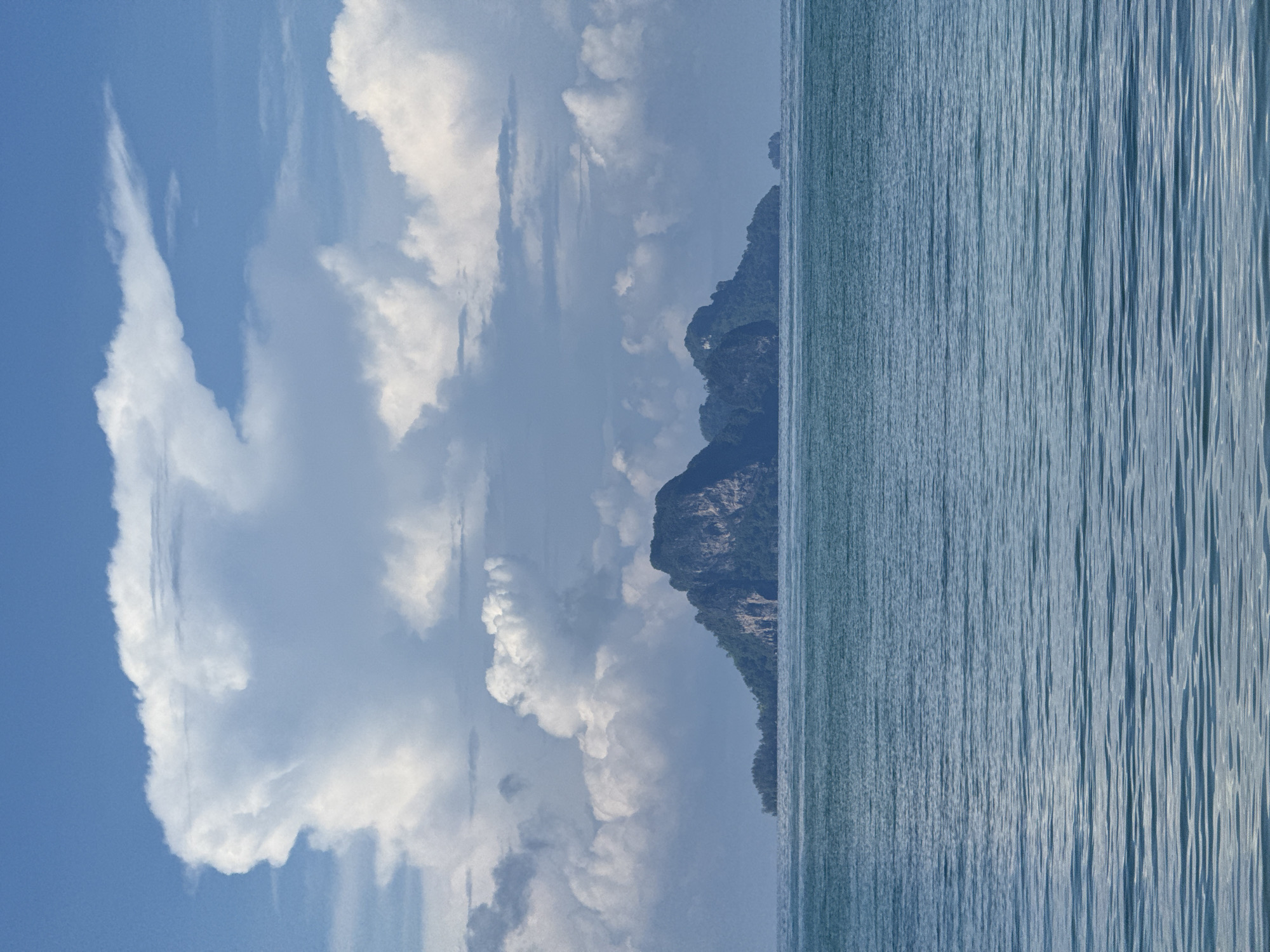 The image depicts a serene seascape with a prominent island in the distance. The sky is a vibrant blue, adorned with large, fluffy white clouds that create dramatic shadows. In the foreground, the ocean is calm, with gentle ripples reflecting the sky. The island, covered in lush green vegetation, rises steeply from the water, its rocky slopes partially obscured by trees. The overall scene suggests a peaceful, natural environment. The location is identified as Ban Klong Haeng, Thailand, known for its picturesque islands and coastal landscapes.