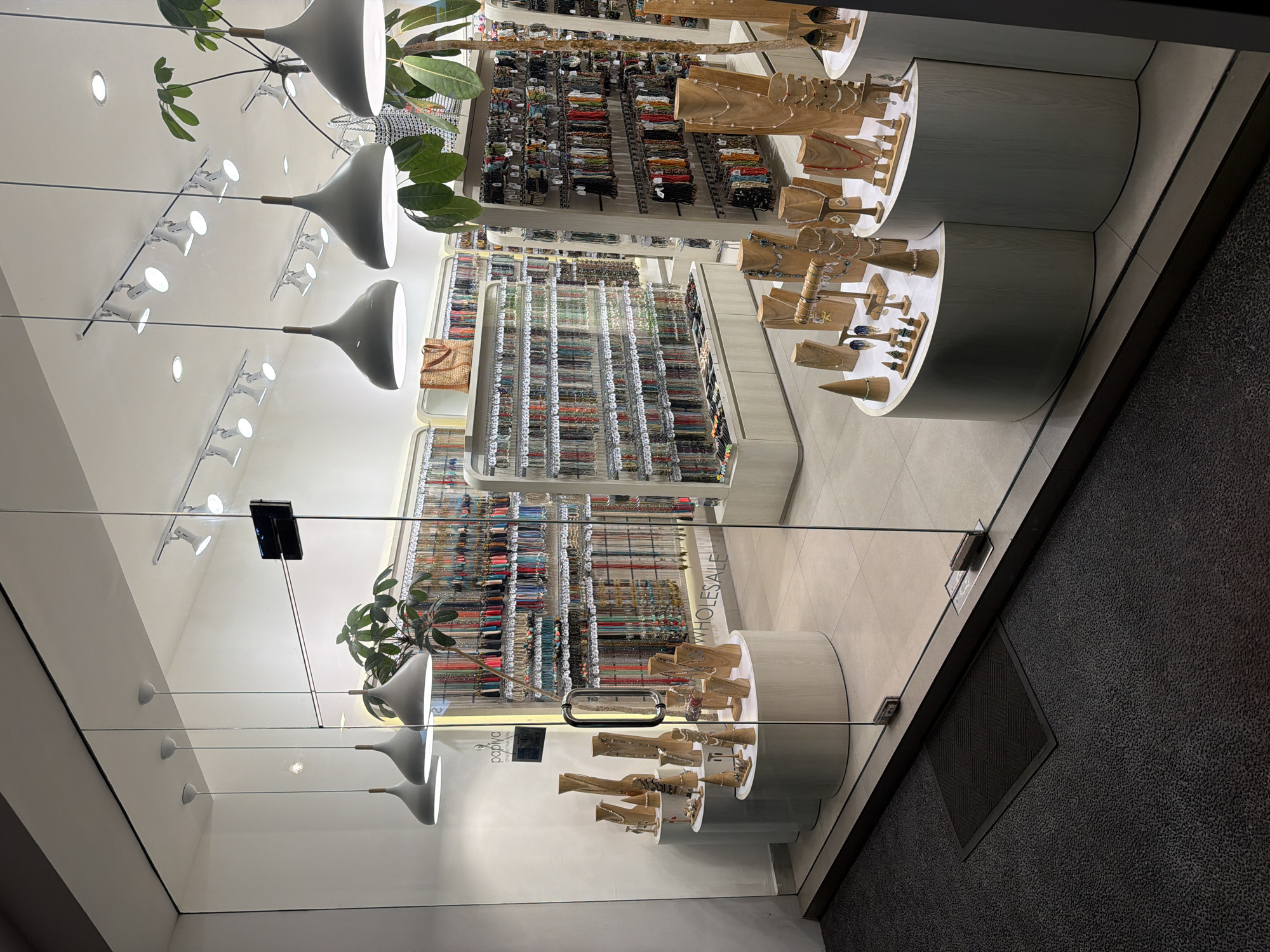 The image is a view of a jewelry store located in Kuta, Indonesia, as seen through a glass storefront. The store displays a wide variety of beads, necklaces, earrings, and other accessories. The jewelry is arranged on shelves, with some items showcased on wooden stands. The interior of the store is well-lit with overhead lighting fixtures. There are also plants inside the store, adding to the decor. The store's name is not visible, but a sign with the word "WHOLESALE" is partly visible. The background is a neutral color.