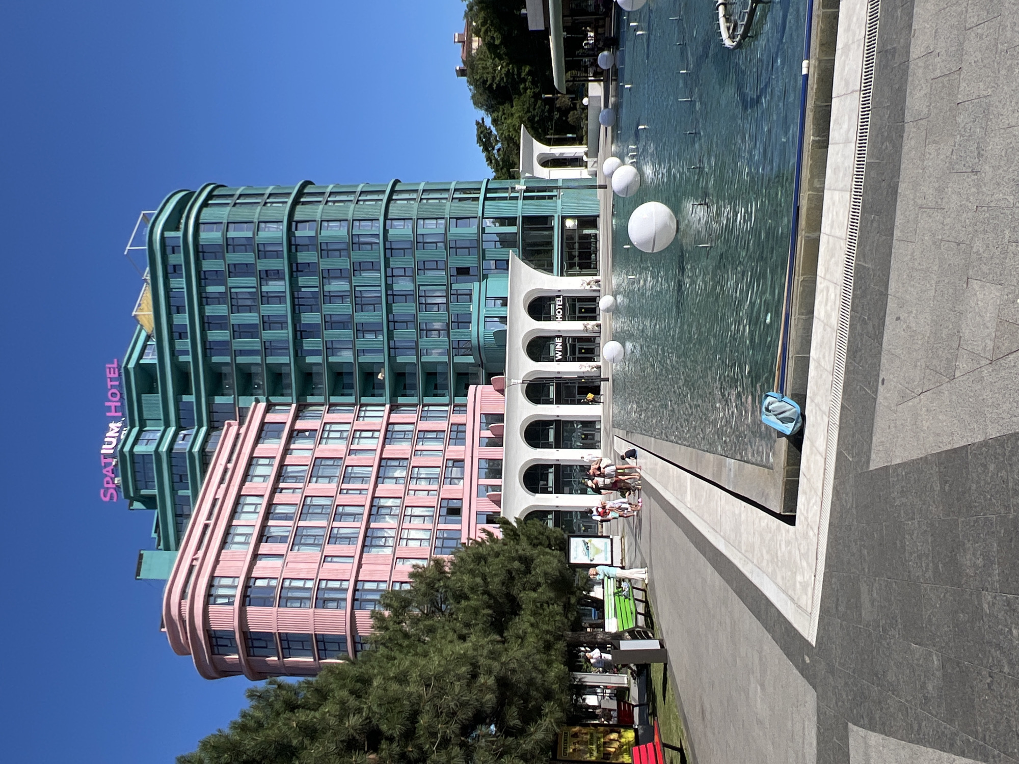 The image captures a bright, sunny day in Odesa, Ukraine, with a clear blue sky. The scene features two strikingly modern hotels, one in a vibrant pink and the other in a teal green, standing tall against the sky. The pink hotel's facade is adorned with numerous windows arranged in a grid, while the teal hotel exhibits a more curved, architectural design with a similar window pattern.
In the foreground, a large fountain area is visible, with several white spherical objects floating on the water's surface. The water reflects the surrounding buildings and the bright sky, creating a dynamic visual. A tiled walkway runs alongside the fountain, bordered by grey stone paving.
A group of people is seen strolling along the walkway near the fountain, enjoying the pleasant weather. Some individuals are seated on a green bench, and others are standing, engaged in conversation or simply observing the surroundings. The presence of trees suggests a park-like setting or a landscaped area adjacent to the hotels and fountain.
Visible text on the pink hotel reads "SPATIUM HOTEL" in large, pink, capitalized letters. Another sign near the arched entrance of one of the buildings displays the word "HOTEL" in white lettering above "WINE". The overall atmosphere is one of a lively urban environment on a beautiful summer day.
