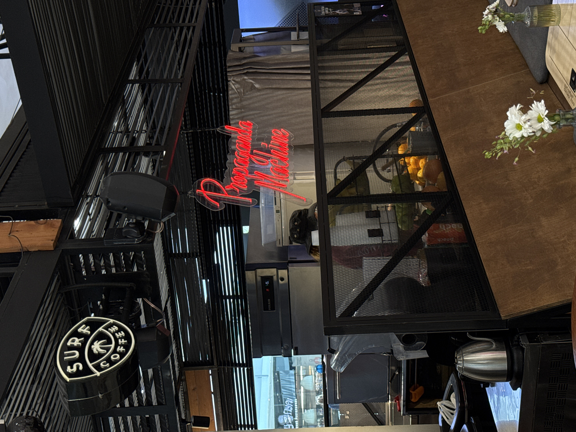 This image captures the interior of a Surf Coffee establishment located in Sochi, Russia, likely within a shopping center such as MoreMall. The scene presents an industrial-modern aesthetic with predominant black and brown tones.
Suspended from the black metal slatted ceiling structure is an illuminated, circular black sign displaying "SURF COFFEE" in white capital letters, centered with a white palm tree silhouette. Adjacent to it, a red neon sign in cursive script spells out "Propaganda Machine." The ceiling and upper structural elements consist of black metal slats and a grid pattern.
In the mid-ground, a black metal mesh railing with diagonal bracing forms a partition, separating the customer area from an operational or service space. Behind this railing, a commercial-grade stainless steel refrigerator or freezer unit, marked "POLAIR," displays a red digital reading of "2:15." Other kitchen or bar equipment, including an obscured counter, is visible. A person wearing a black bucket hat is partially visible behind the equipment. A grey curtain or divider is situated in the background of this service area. A basket filled with oranges is also visible behind the mesh railing.
In the foreground, an electric silver kettle rests on a counter surface on the left. To the right, part of a seating booth is visible, featuring a light brown wooden panel backrest and a dark grey upholstered seat cushion. A light-colored wooden table supports two small clear glass vases, each containing a small bouquet of white daisy-like flowers with green foliage.
Through a large opening or window in the background, a reflective view of an adjacent indoor commercial area is present. Partially legible Cyrillic text, including "ГУРИ" and "ресторан" (restaurant), is visible on a distant sign, suggesting the cafe is situated within a larger commercial complex. The lighting is ambient, supplemented by the illuminated signs.