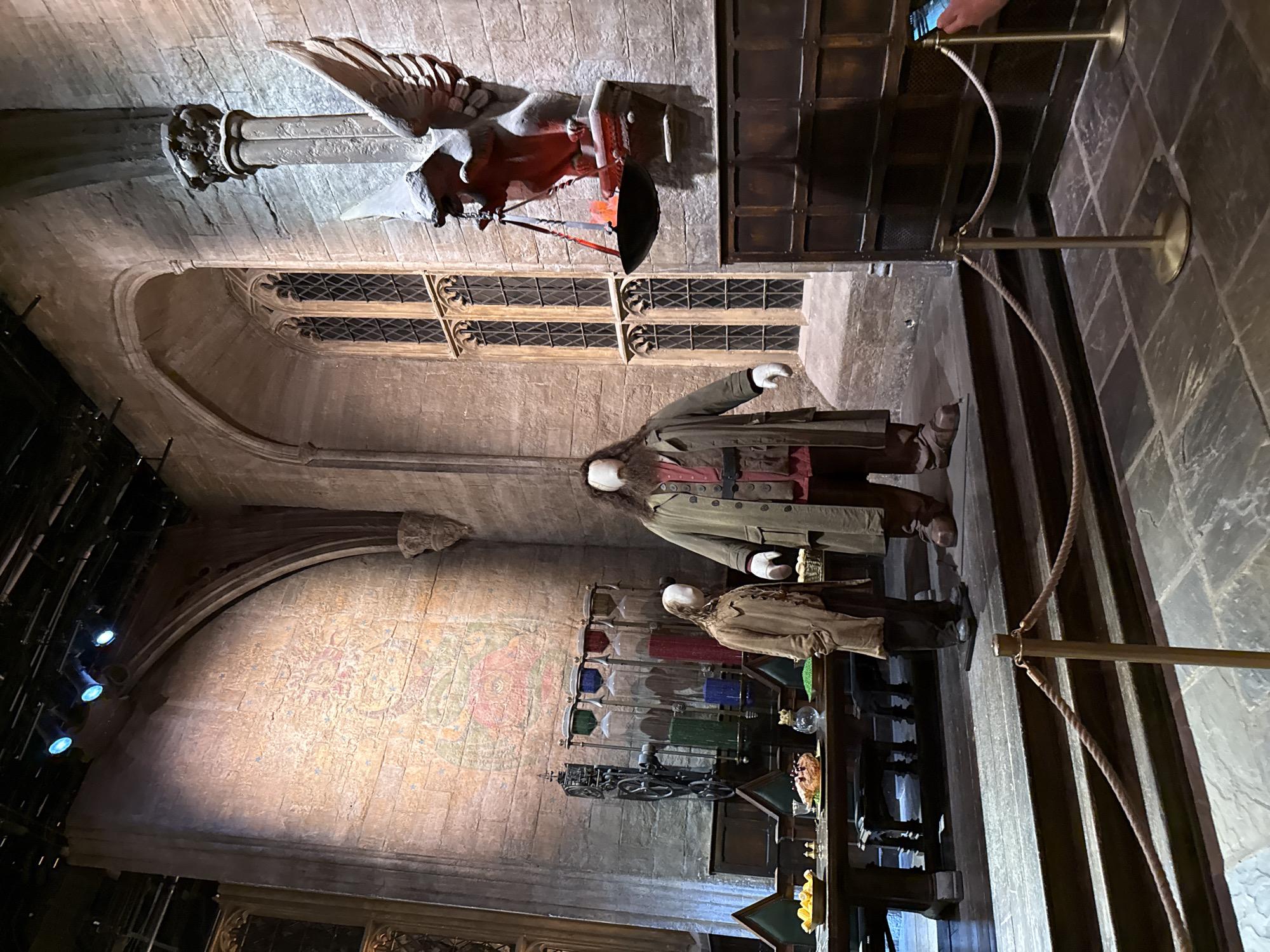 This is a photograph of a meticulously recreated Hogwarts Great Hall set, located at Warner Bros. Studio Tour London – The Making of Harry Potter in Watford, United Kingdom. The scene features two mannequins, one depicting Hagrid in his signature long coat and another of a young boy in a trench coat. They stand on a raised wooden platform in front of a long, imposing wooden table laden with food props, including a roast chicken and bowls of fruit. Behind the table, a series of hourglasses filled with colored liquids are displayed, suggesting potion-making materials.
The backdrop is a grand stone wall with a large, arched window characteristic of medieval architecture. High on the wall, a stone griffin statue is mounted, holding a suspended cauldron that emits a red glow, creating an atmospheric effect. The lighting is theatrical, with blue spotlights directed at the ceiling, illuminating the upper reaches of the hall. The floor is made of large, grey stone tiles. Ropes and stanchions are visible in the foreground, indicating this is part of a guided tour or exhibit.
