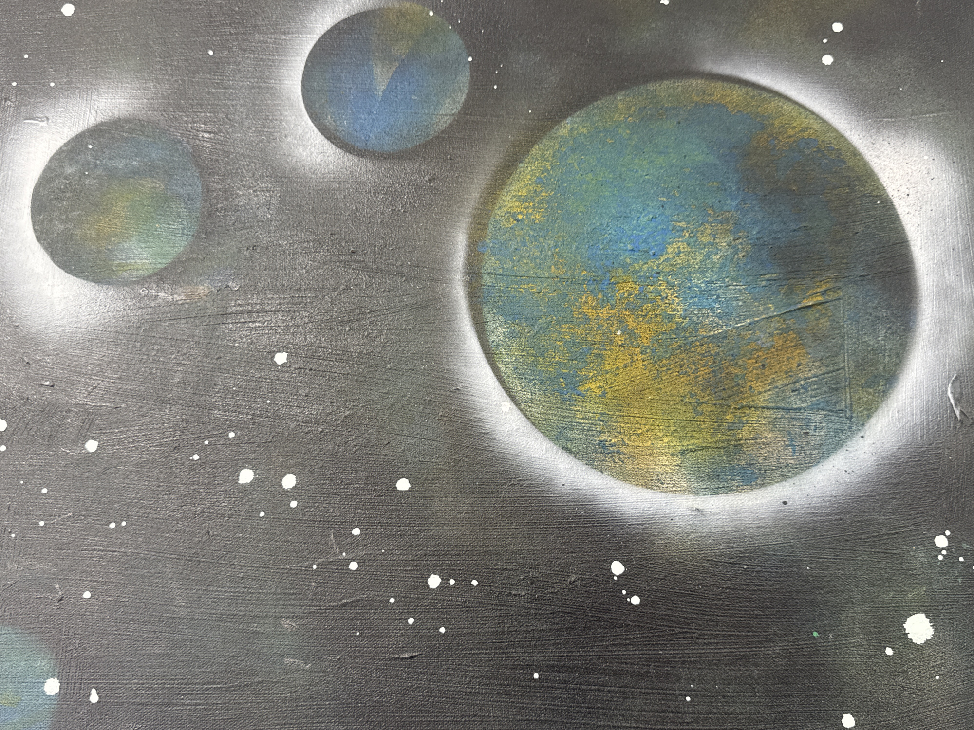 The image depicts a painting of a celestial scene with three spherical objects resembling planets against a dark gray background dotted with white specks, representing stars. The planets are varying sizes and colored in shades of blue, yellow, and green, and have white halos. The painting style has a textured, possibly spray-painted, effect. There is no evidence suggesting this is physically located in Netanya, Israel.