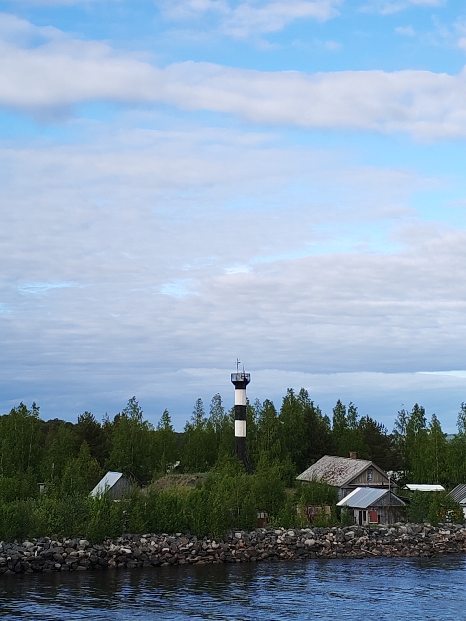 The image shows a view of Ozernoe Ust'e, Russia. A black and white striped lighthouse stands amid green trees. Several rustic wooden houses are nestled near the rocky shoreline of a body of water, likely Lake Ladoga. The sky is mostly covered with white clouds.