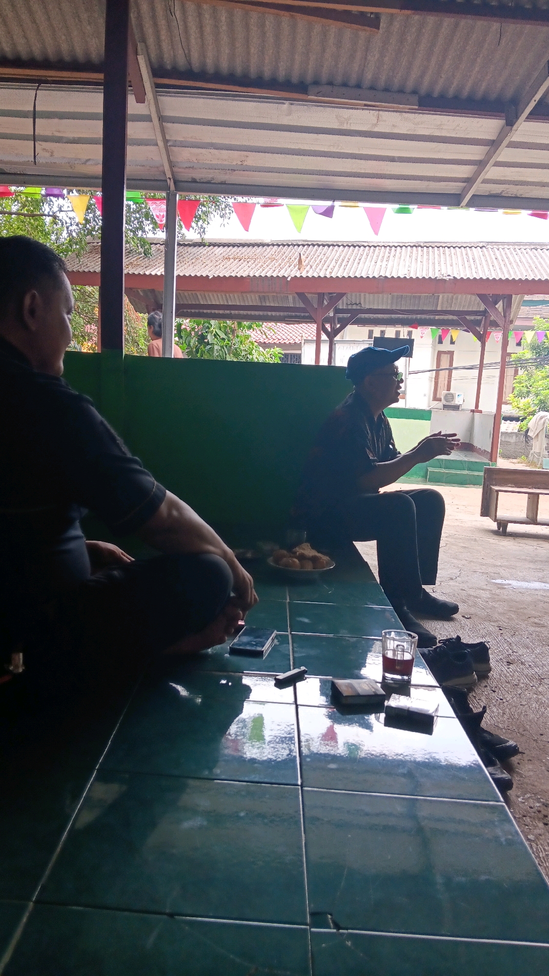 The image depicts a casual gathering of two men sitting on a tiled surface under a corrugated metal roof, likely at an outdoor cafe or eatery in Depok, Indonesia. The man on the left is seated cross-legged, wearing a dark shirt and pants, with his back partially to the camera. The man on the right is seated with his legs extended, wearing a dark shirt, pants, and a blue cap. He appears to be gesturing with his hands as if in conversation. On the tiled surface in front of them are a plate of snacks, a glass of red liquid, a smartphone, and what appear to be a lighter and small packets. In the background, there is a green wall, trees, and parts of buildings. Colorful triangular flags are strung across the top of the scene, suggesting a festive atmosphere. The overall impression is of a relaxed social interaction in a common, open-air setting.