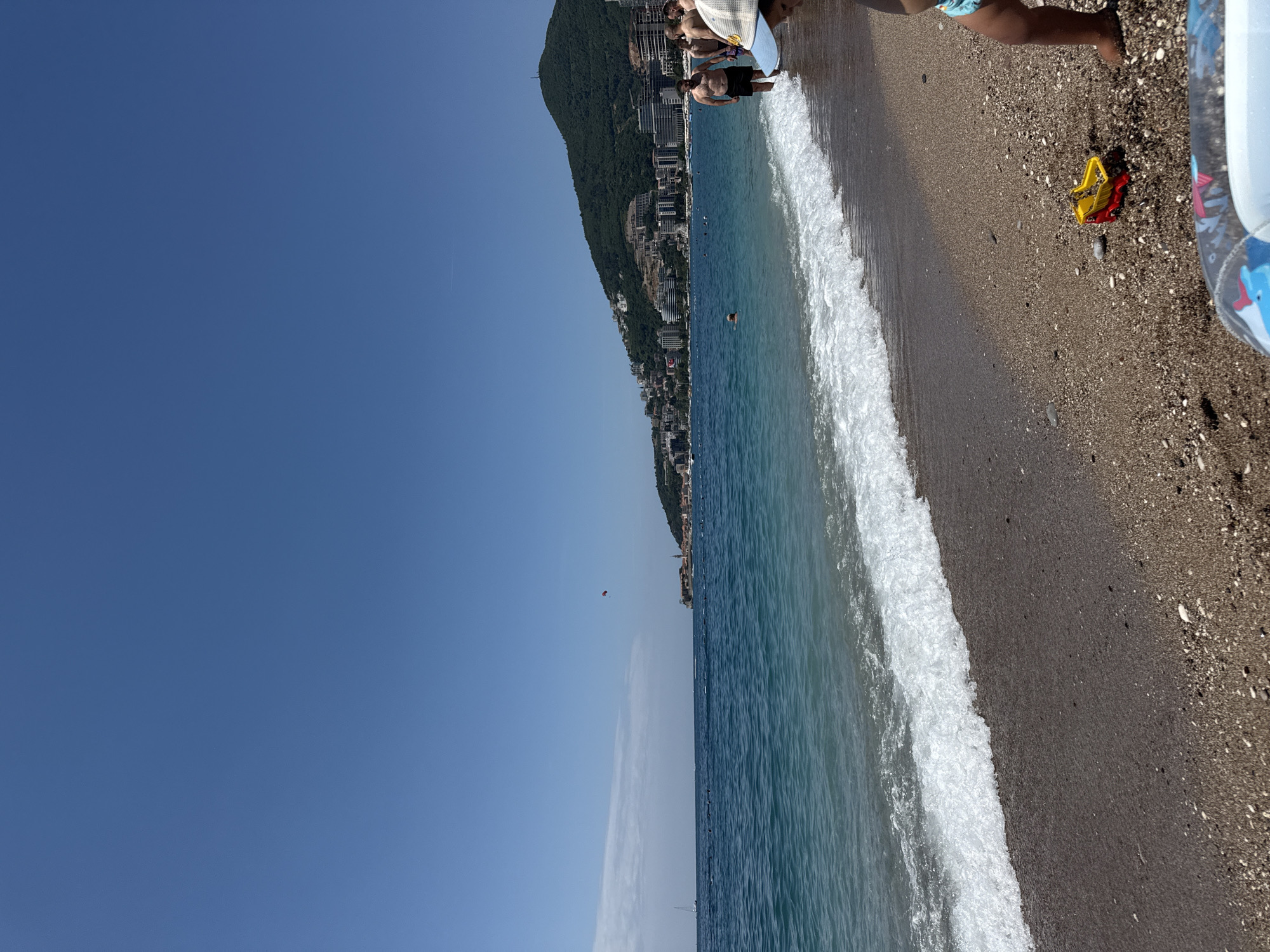 This image captures a vibrant summer day on a pebbly beach in Budva, Montenegro. The scene is dominated by a clear, bright blue sky that stretches across the top and left, with a faint airplane contrail visible. Below the sky, the serene blue-green waters of the Adriatic Sea extend, showing gentle white waves breaking along the shore.
In the mid-ground, a long, verdant mountain range slopes down towards the sea, densely dotted with numerous buildings and resorts, indicating a thriving coastal town. Within the sea, a lone swimmer is visible. High above the water in the distance, a person is parasailing, identifiable by a distinct red parachute, adding to the leisurely atmosphere.
On the immediate right foreground, the pebbly beach is clearly visible, showing the legs of a person, likely a child, near the water's edge. A bright yellow and red beach toy, possibly a bucket or shovel, rests on the sand, alongside a partial view of an inflatable ring or float. Further back on the beach, other individuals are present; one is partially obscured, possibly resting, while another person stands. The bright, natural light suggests it is daytime, likely mid-morning to afternoon. The overall scene conveys a sense of summer vacation, relaxation, and outdoor activity. No visible text is present in the image.
