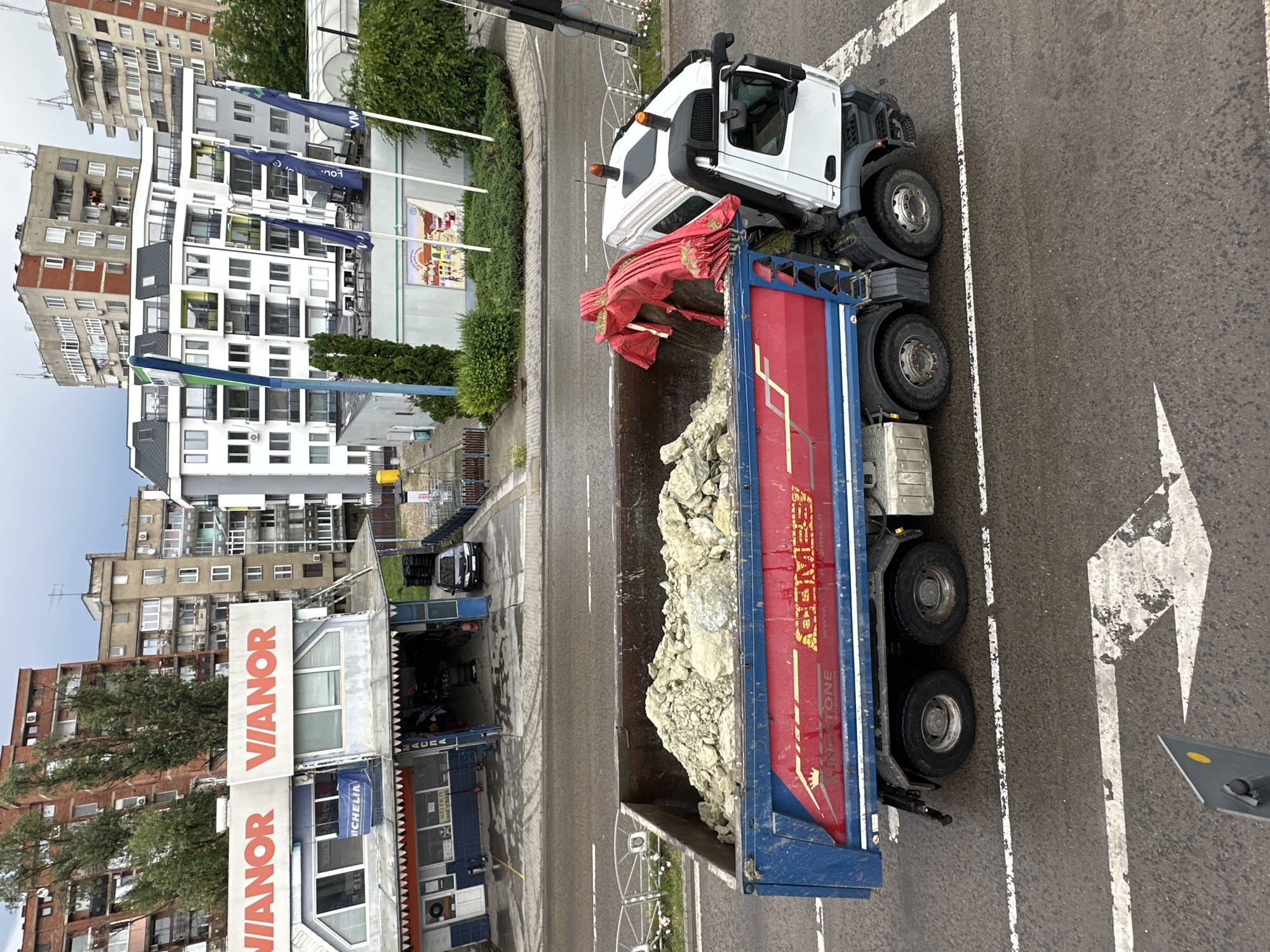 The image provides an elevated view of a city street in Veliko Tarnovo, Bulgaria, captured during an overcast day. In the foreground, a white and blue dump truck is stationary on the asphalt road, its bed loaded with light-colored rocks or construction aggregate. A red tarp is gathered at the front of the dump bed. The truck displays branding with "ACMESI" in yellow text, "KINGSTONE" in white, and the website "www.acmesi.com".
The road features white lane markings, including a dashed line and a prominent white arrow indicating a turn or direction. Across the street from the truck, a commercial strip includes a "VIANOR" tire service center, identified by its large red and white signage, and a smaller blue sign for "MICHELIN". A black car is parked near the service center.
Behind the commercial establishments, various multi-story residential or mixed-use buildings line the street, some featuring balconies and air conditioning units. Green trees and bushes are scattered throughout the scene. Several blue flags with partially visible text, such as "FOU..." and "VIA", are flown near one of the buildings. The overall scene depicts an active urban environment with elements of commerce, residential living, and industrial transport. No people are explicitly visible.