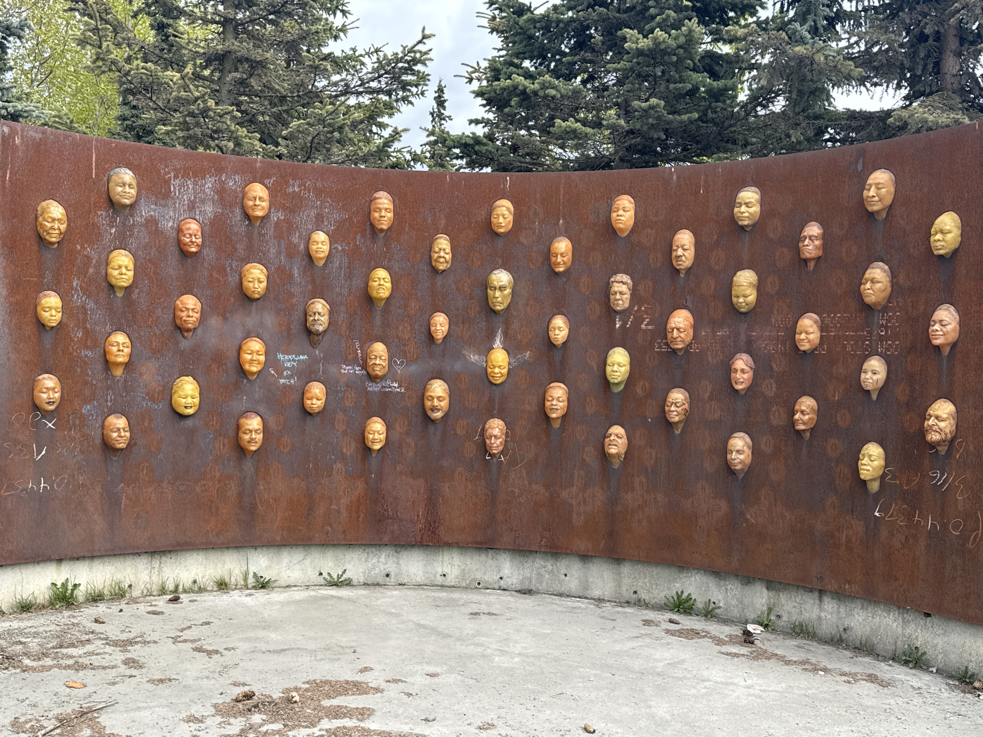 The image shows a curved wall of rusted metal with dozens of human faces protruding from it. The faces are made of a material that appears to be bronze or a similar metal, and they are arranged in a grid-like pattern. The wall is set on a concrete base, and there is some green vegetation growing nearby. The background features green trees. Based on the style of the artwork and the inscription on the wall, it can be deduced this location is the Delaney Park Strip in Anchorage, Alaska, United States of America.