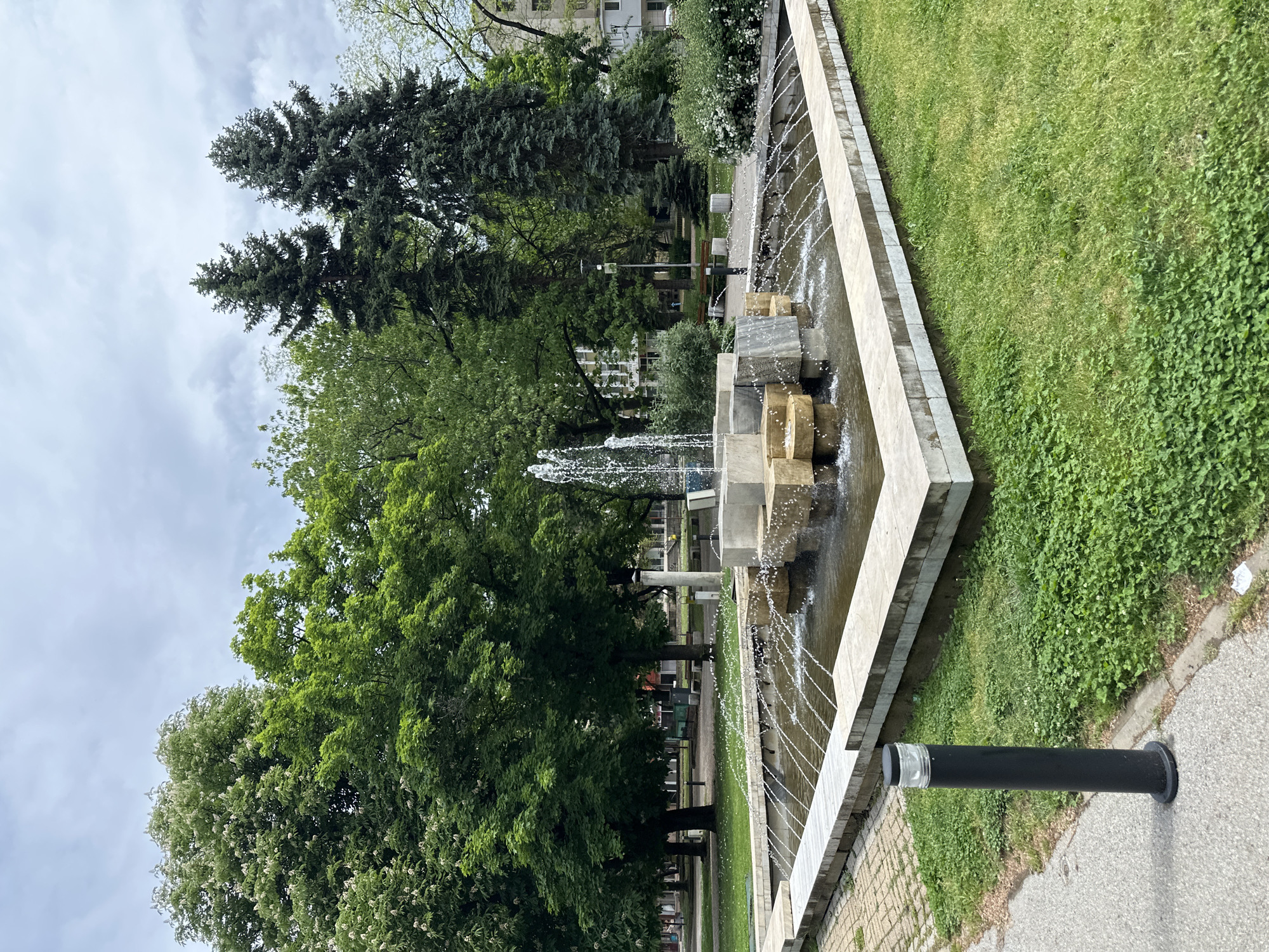 A park in Montana, Bulgaria, contains a rectangular concrete fountain with blocky geometric stone structures in the center, where water spouts upwards and cascades down the sides. The fountain is flanked by lush green grass on one side and a paved pathway on the other. Beyond the fountain, dense foliage of trees and shrubs lines the background, with a multi-story building partially visible through the trees. The sky is overcast with grey clouds. A black, cylindrical bollard stands near the pathway.