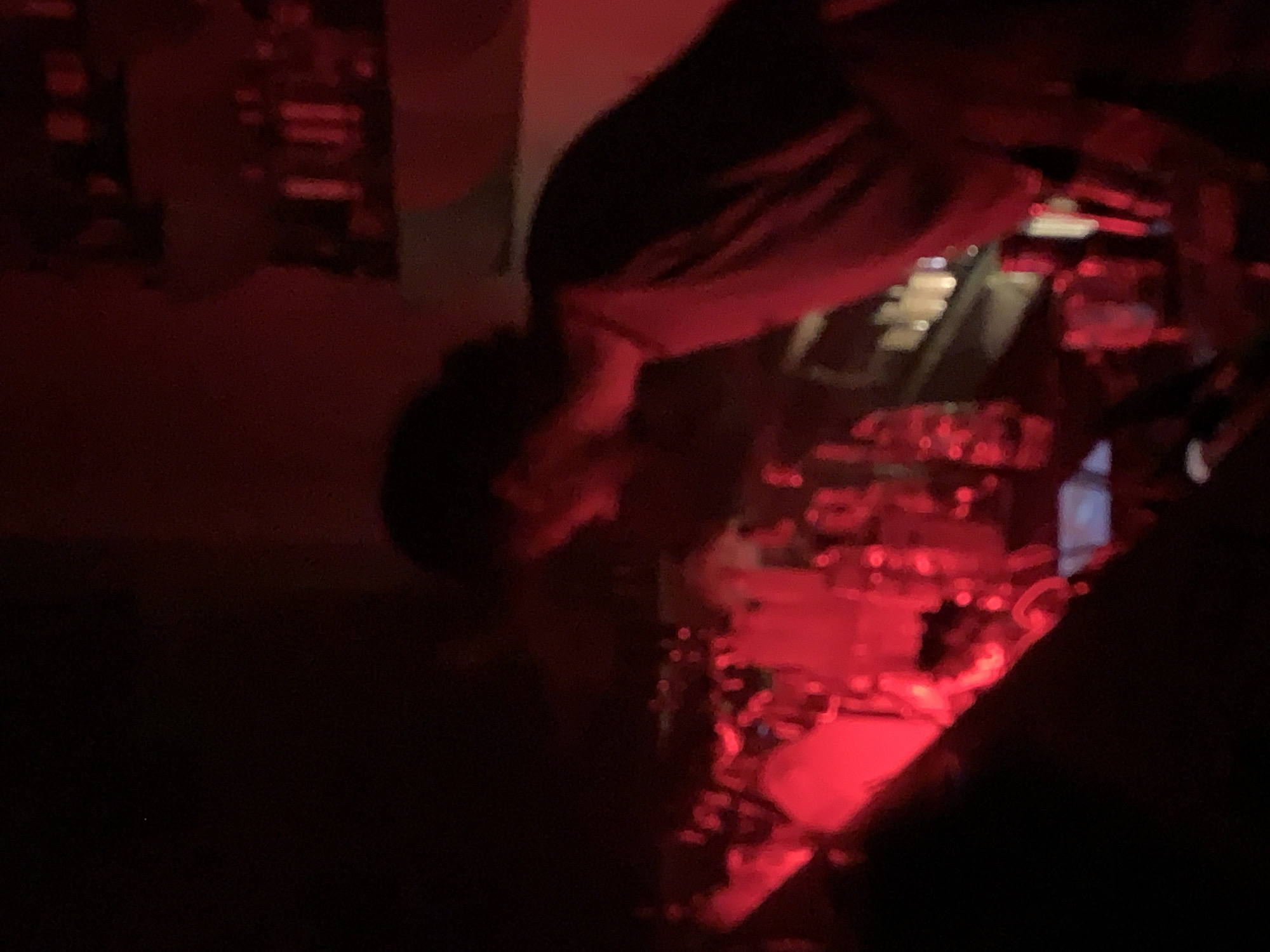 The image shows a person with dark hair bent over a drum set under intense red lighting. The background is dark and indistinct, but appears to be a wall. The location is possibly Tel-Aviv, Israel, but without further information, this is not confirmed.
