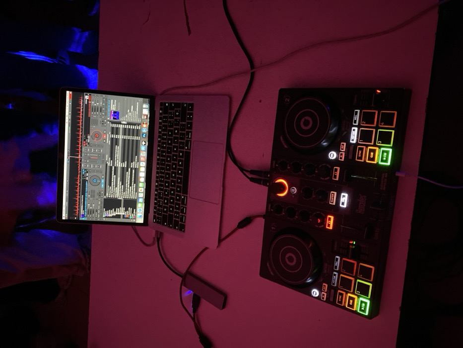 An electronic music setup is positioned on a light-colored, possibly white, flat surface in an indoor location in Ushuaia, Argentina. The scene is primarily illuminated by ambient pink light, with a blue light source visible in the dark, blurred background. A silver MacBook laptop is open, displaying DJ software with two virtual decks, audio waveforms, and a track list. Directly in front of the laptop, a black Hercules DJControl Inpulse 200 controller is powered on, indicated by various illuminated buttons and pads in green, orange, and white. A silver rectangular USB hub is connected to the laptop's left side via a black cable, and another black cable extends from this hub to the DJ controller. Additional black and a thin white cable are visible, suggesting audio and power connections. No identifiable individuals are clearly present; an indistinct, blurred form is visible in the background under the blue light.