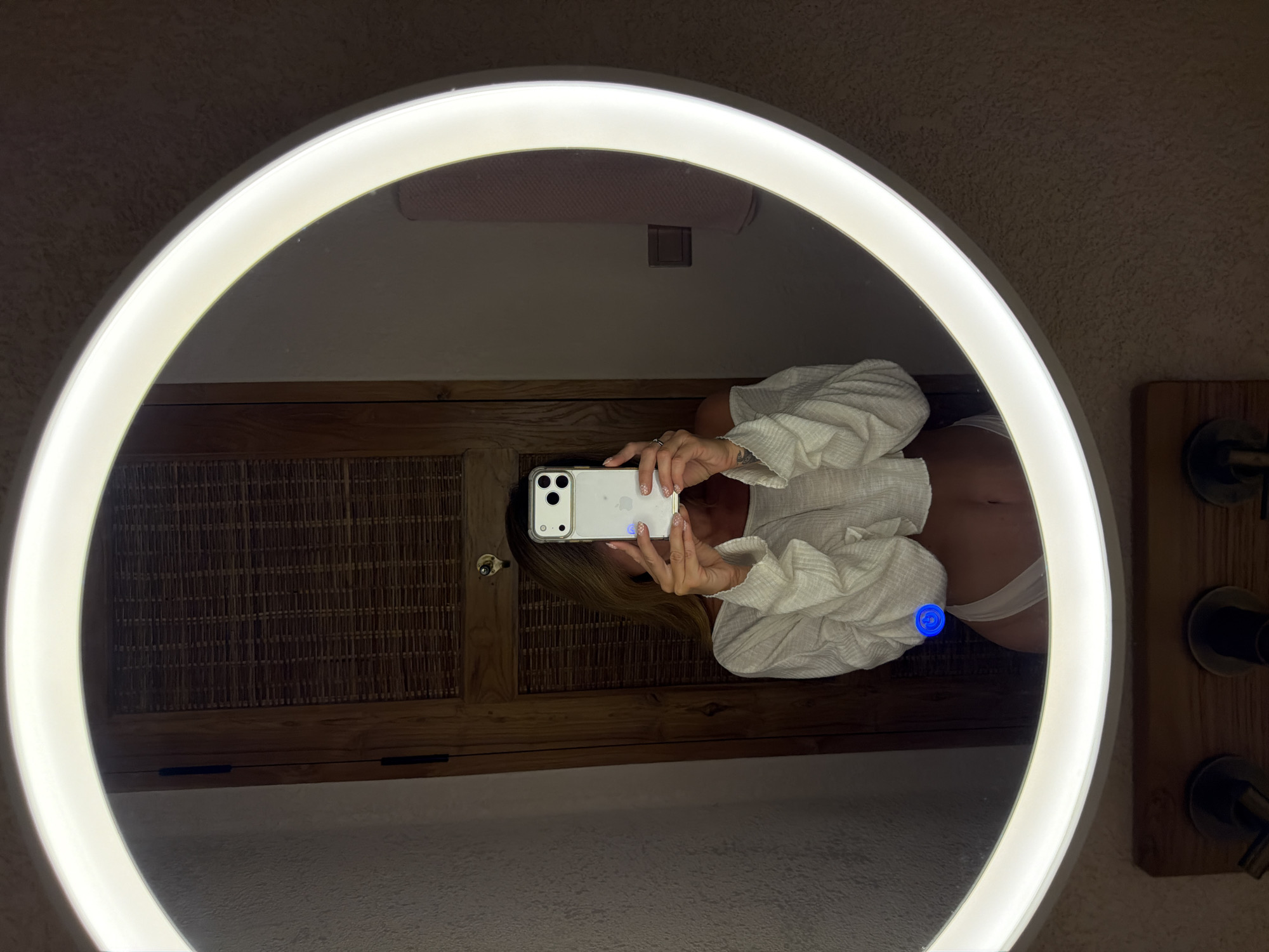 A full-body selfie is taken in a circular mirror with a built-in LED light. The person in the mirror, who appears to be female with blonde hair, is wearing a white cropped long-sleeved top and white underwear. She is holding a white smartphone with a clear case, which is partially obscuring her face. The background reflected in the mirror shows a wooden door with a woven texture, a textured wall, and a light pink towel hanging on a rack. The mirror is positioned above a wooden shelf holding two dark-colored faucets. A blue light is visible on the mirror's surface. The scene appears to be a bathroom. The location is in the city of Mengwi, Indonesia.