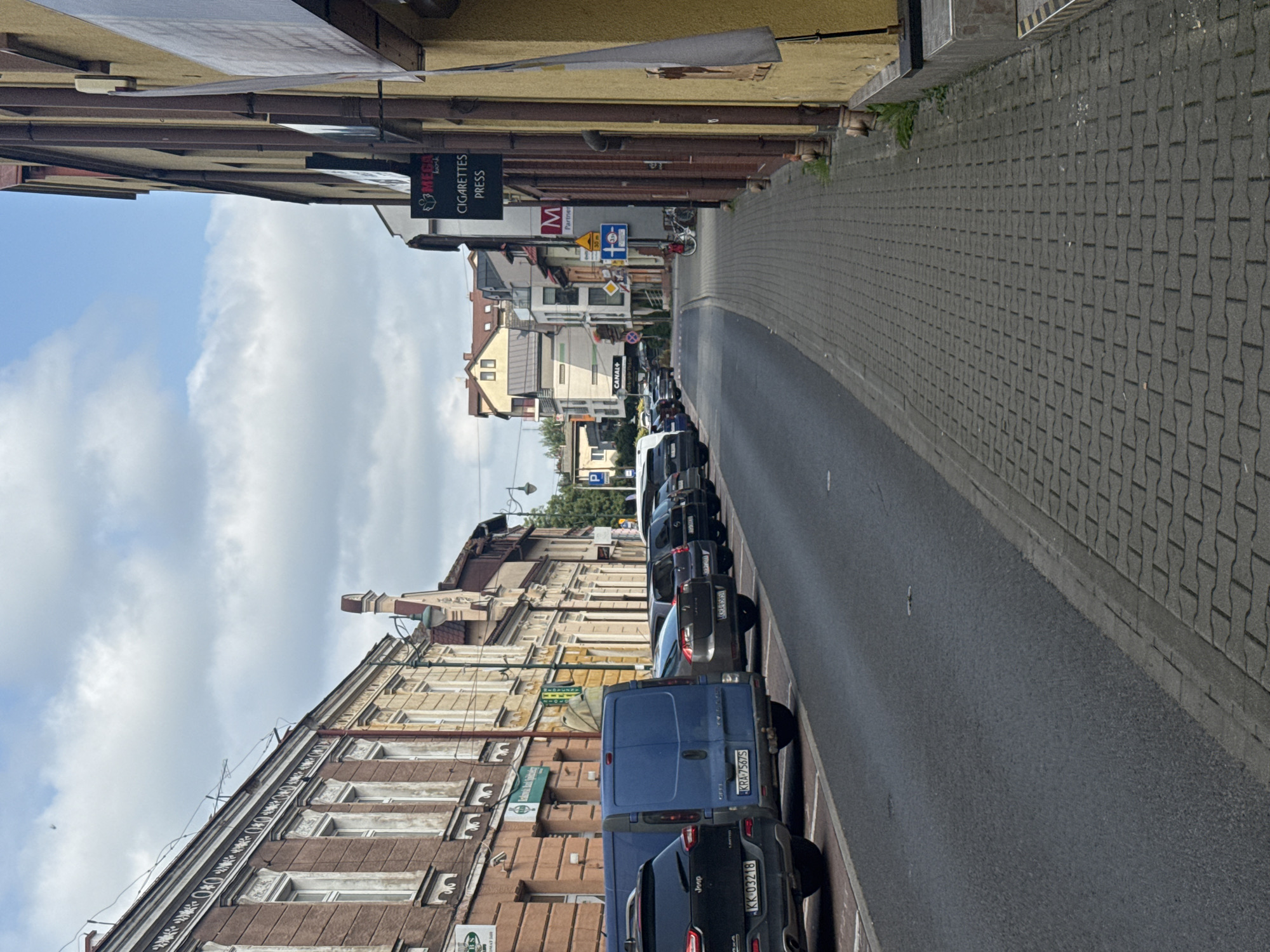 The image depicts a street scene in a city. On the left side of the street, there is a row of parked vehicles, including a black Jeep, a blue van, and several sedans. The buildings lining the street on the left are multi-story structures with architectural details. On the right side of the street, a yellow building with brown trim is prominent, and there are signs advertising "MEGA" and "CIGARETTES PRESS." Further down the street, more buildings are visible, along with streetlights and a bicycle. The street itself is paved, and there is a cobblestone sidewalk on the right. The sky is cloudy.