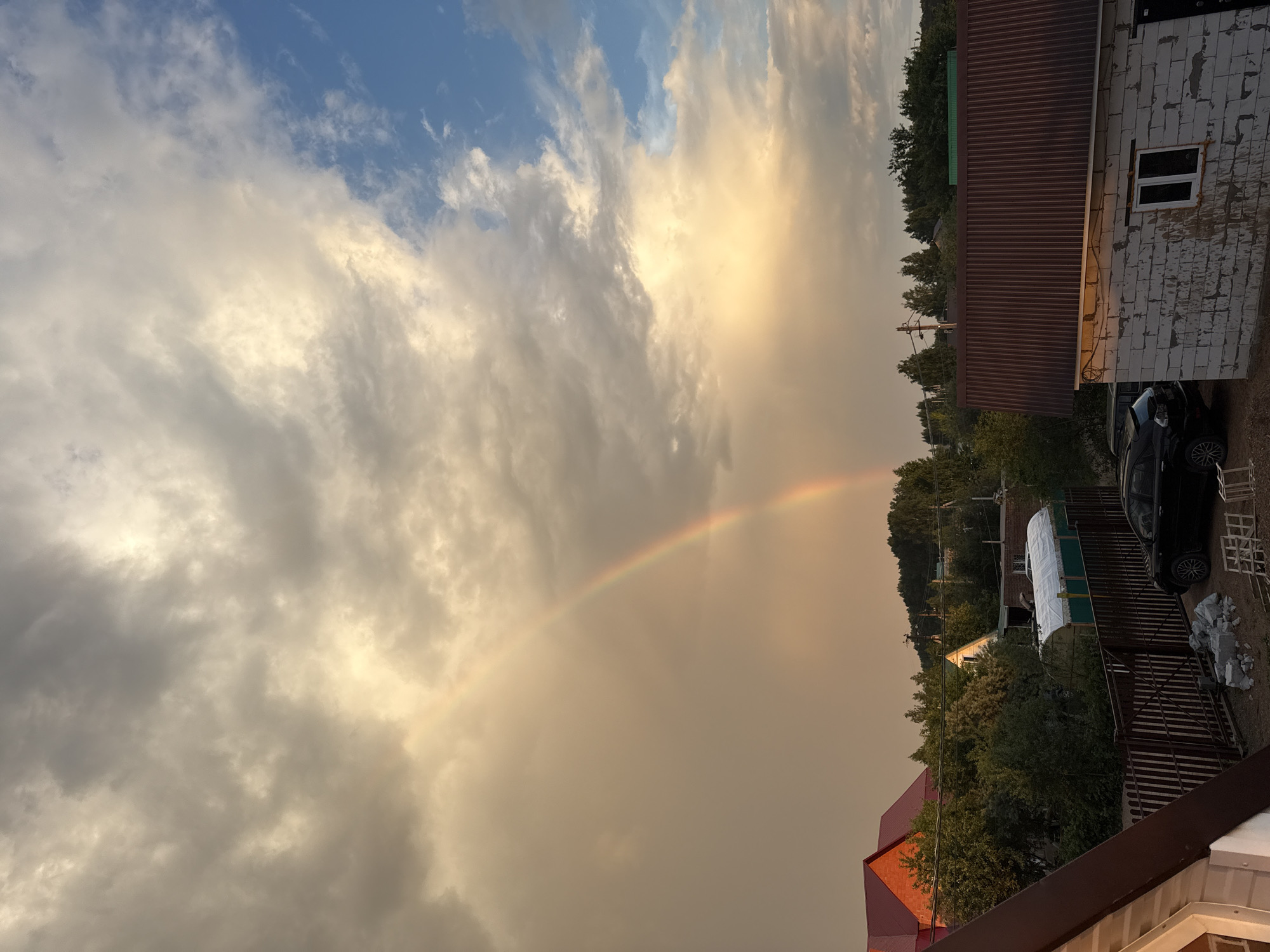 The image depicts a suburban or rural scene during what appears to be sunset or sunrise. A prominent rainbow arcs across a cloudy sky, with patches of blue visible. In the foreground, the roof of a house with a dark brown, corrugated metal roof is visible. To the left, another house with a red, pitched roof is partially visible. A dark-colored car is parked in front of a wooden fence. Beyond the fence, there are trees and several other buildings, including a white structure covered with a tarp and a brick building with a dark corrugated roof and a window. Electrical poles and wires are also present. The overall lighting suggests diffused daylight, possibly after or during a rain shower.