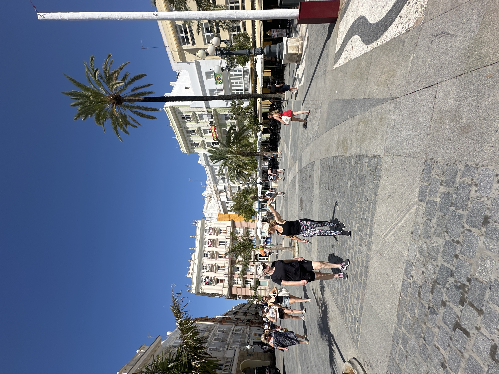 The image depicts a sun-drenched plaza in Cádiz, Spain, likely the Plaza de las Flores, given its characteristic architecture and palm trees. Several pedestrians are walking across the wide, paved plaza. In the foreground, two women are walking towards the camera; one wears a black tank top and patterned leggings, the other a black t-shirt and shorts. Other individuals are visible in the mid-ground, some walking, some standing. The background features multi-story buildings with balconies and awnings, common in Andalusian cities, with palm trees interspersed among them. A tall white pole, possibly a flagpole, is visible on the right side of the frame. The sky is a clear, bright blue. The pavement is a mix of granite cobblestones and larger stone slabs, with a decorative black and white pattern on the right side. The overall scene conveys a lively, daytime atmosphere in a European city.