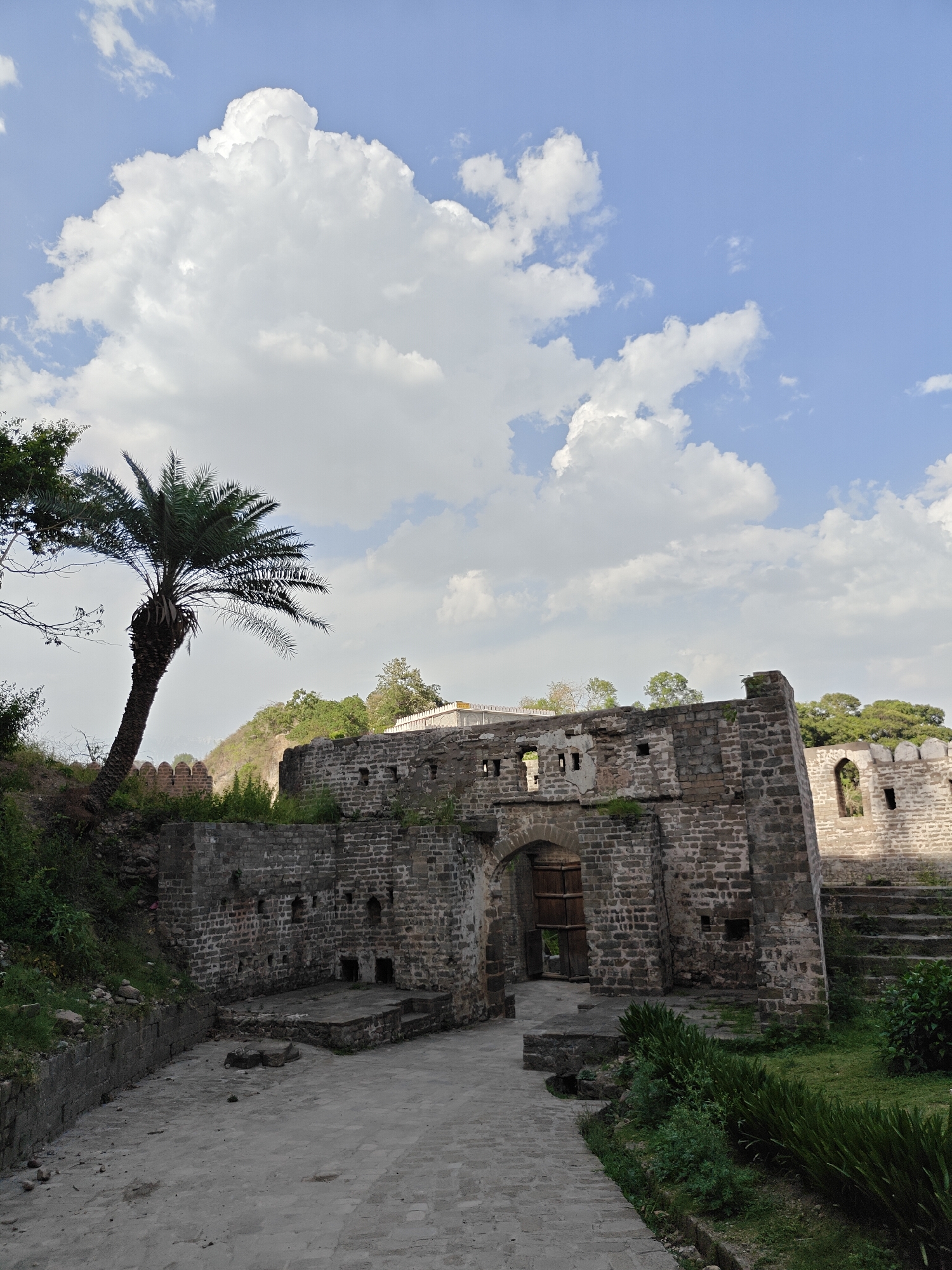 The image shows a historic stone structure in Kaswara, India, under a partly cloudy blue sky. The structure features a gateway with large wooden doors slightly ajar, and sections of stone walls with multiple small openings. A palm tree is visible on the left, partially obscuring a grassy hill in the background. The foreground consists of a stone-paved path leading to the gateway, with patches of green vegetation along the sides. Steps are visible on the right. The stone structure appears aged and weathered.