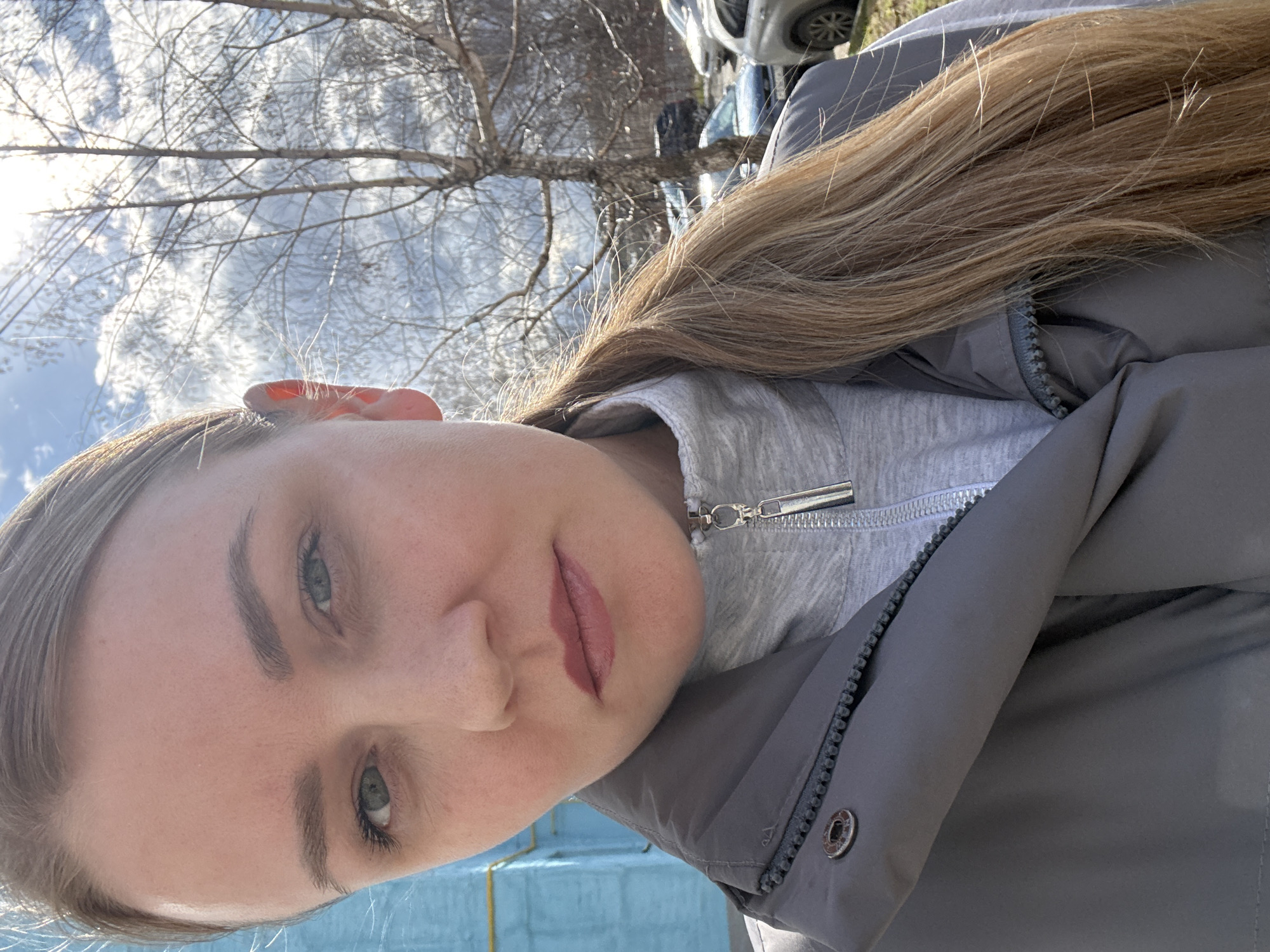A young woman is featured in a close-up, outdoor selfie, looking directly at the camera. She has long, light brown hair that flows over her shoulders. Her light-colored eyes (likely green or blue-green) are visible, framed by groomed eyebrows and light mascara. Her lips are painted with a soft pink or mauve lipstick. She wears a light gray top with a zipped collar, layered under a dark gray jacket that also features a zipper and a visible snap button.
Behind her, bare tree branches reach across a bright, partially cloudy sky, suggesting a colder season like late fall or early spring. Strong sunlight illuminates the scene. In the background, parts of parked cars are visible, alongside a light blue structure, possibly a fence or wall, with a yellow line at its base. The setting appears to be an urban or suburban outdoor area, consistent with Chaikovsky, Russia. The time of day is daytime, under a clear sky, and her expression is neutral and calm. No visible text is present.